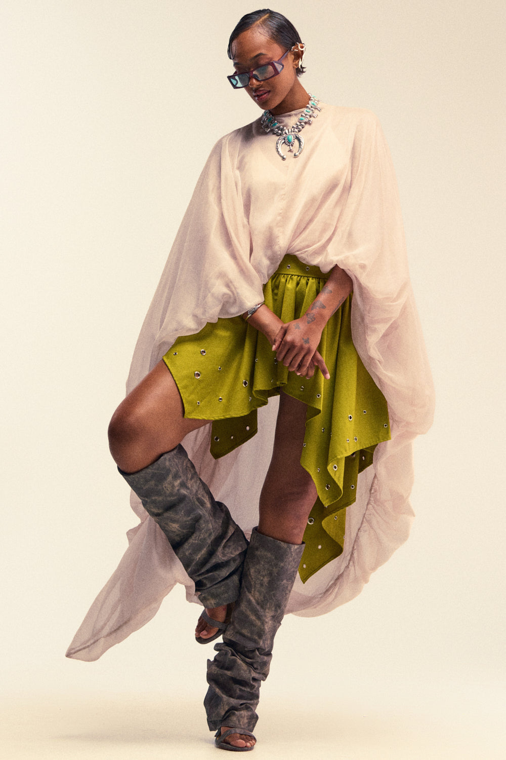 Wearing the HEAD IN THE CLOUDS CHIFFON BLOUSE, this person pairs the pale, sheer draped top with a lime green asymmetrical embellished skirt, gray slouchy boots, a bold statement necklace, and sunglasses while posing confidently against a neutral background.