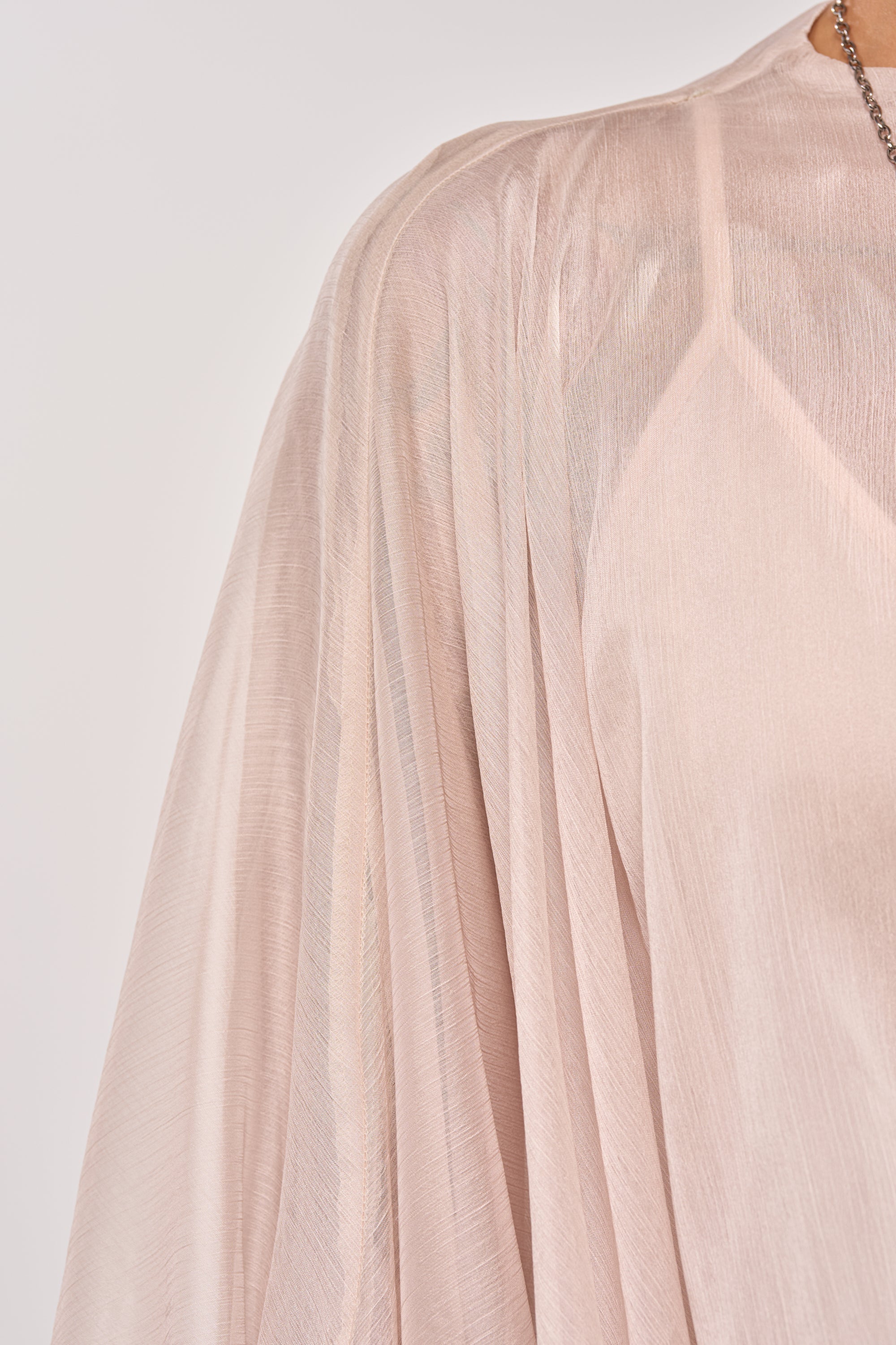 A close-up of the HEAD IN THE CLOUDS CHIFFON BLOUSE in light pink, showing its sheer fabric, soft pleats, and elegant draping over the shoulder, paired with a delicate chain necklace at the neckline. 6