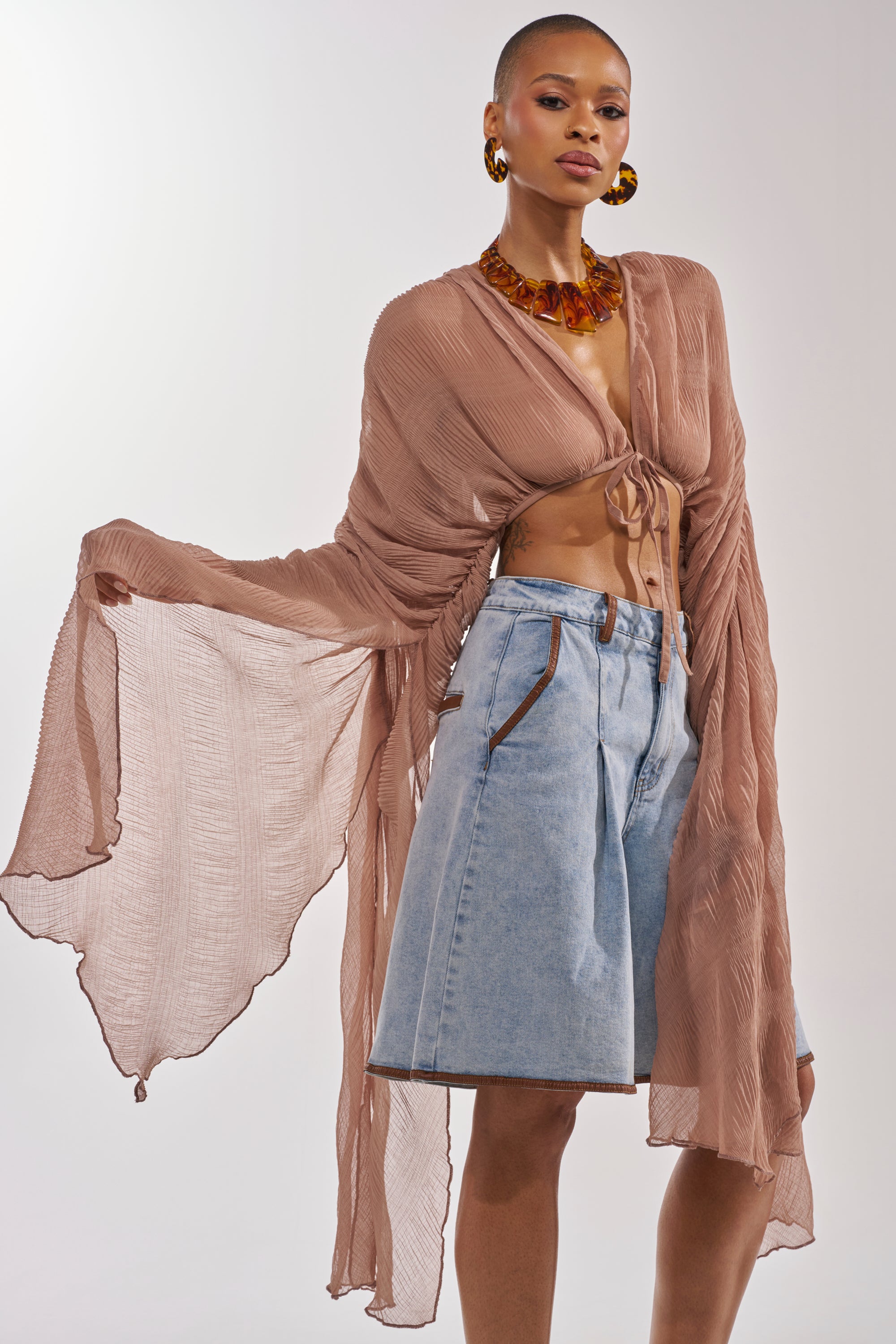 A woman models the FLY AWAY CHIFFON BLOUSE IN TAUPE, featuring sheer fabric, wide sleeves, and a tie front, styled with high-waisted denim shorts. She accessorizes with bold earrings and a chunky necklace against a plain background. 0