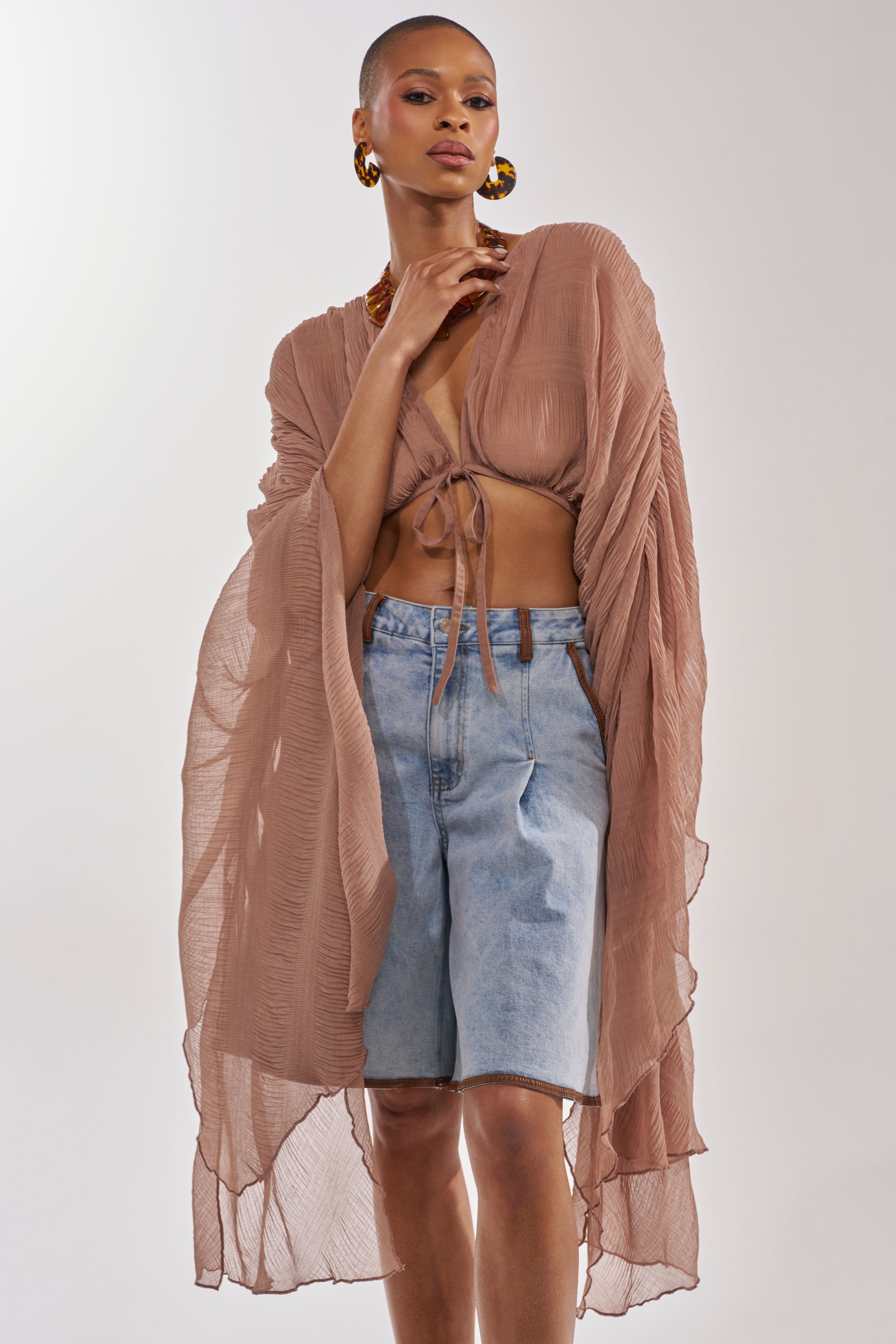 Wearing the FLY AWAY CHIFFON BLOUSE IN TAUPE with wide sleeves, front tie, and a sheer finish, paired with light blue knee-length denim shorts and gold hoop earrings, a person stands confidently against a light background. 1