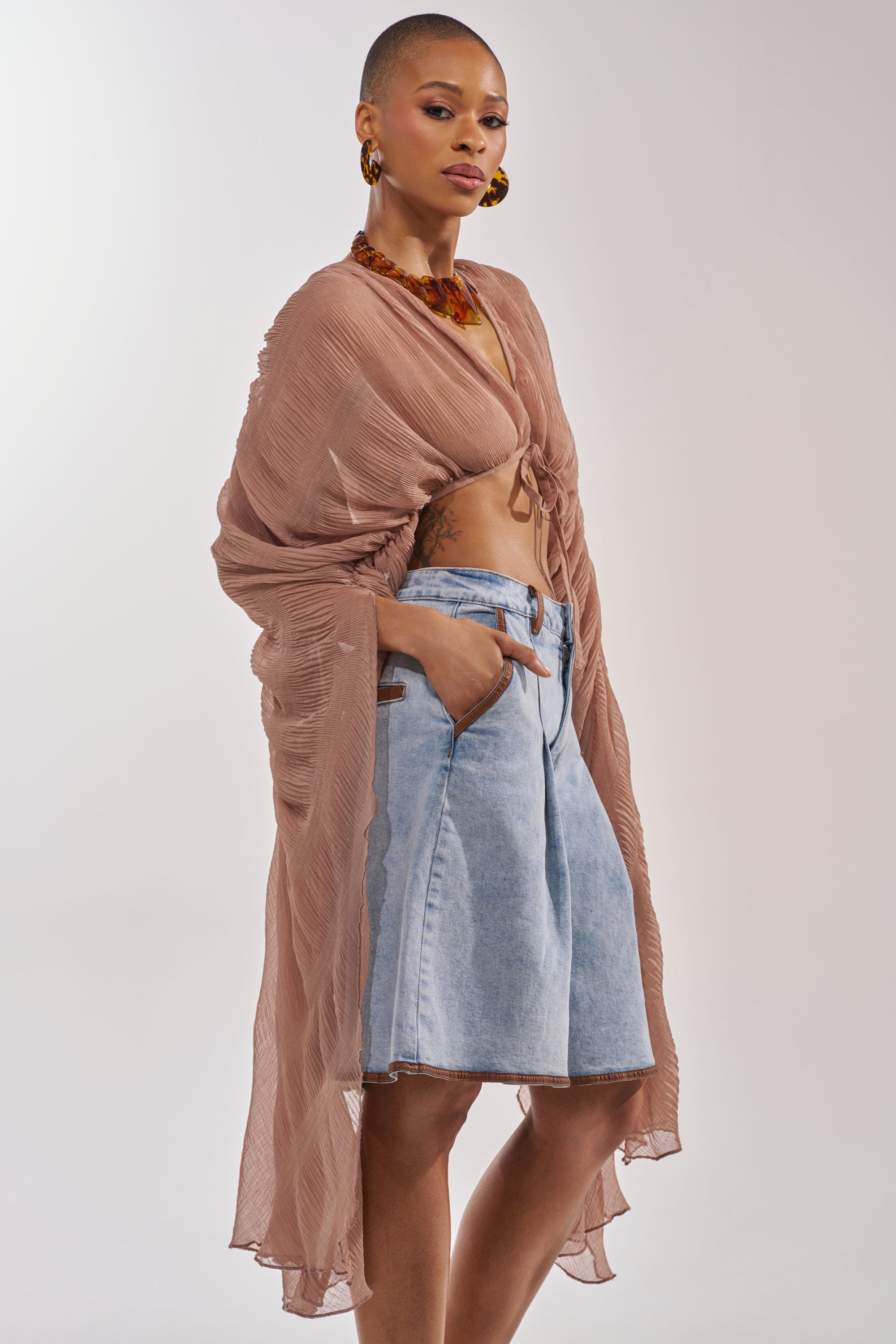 A stylish woman models the FLY AWAY CHIFFON BLOUSE IN TAUPE, tied at the front over a bralette and high-waisted denim shorts, completed with statement earrings and a chunky necklace. 2