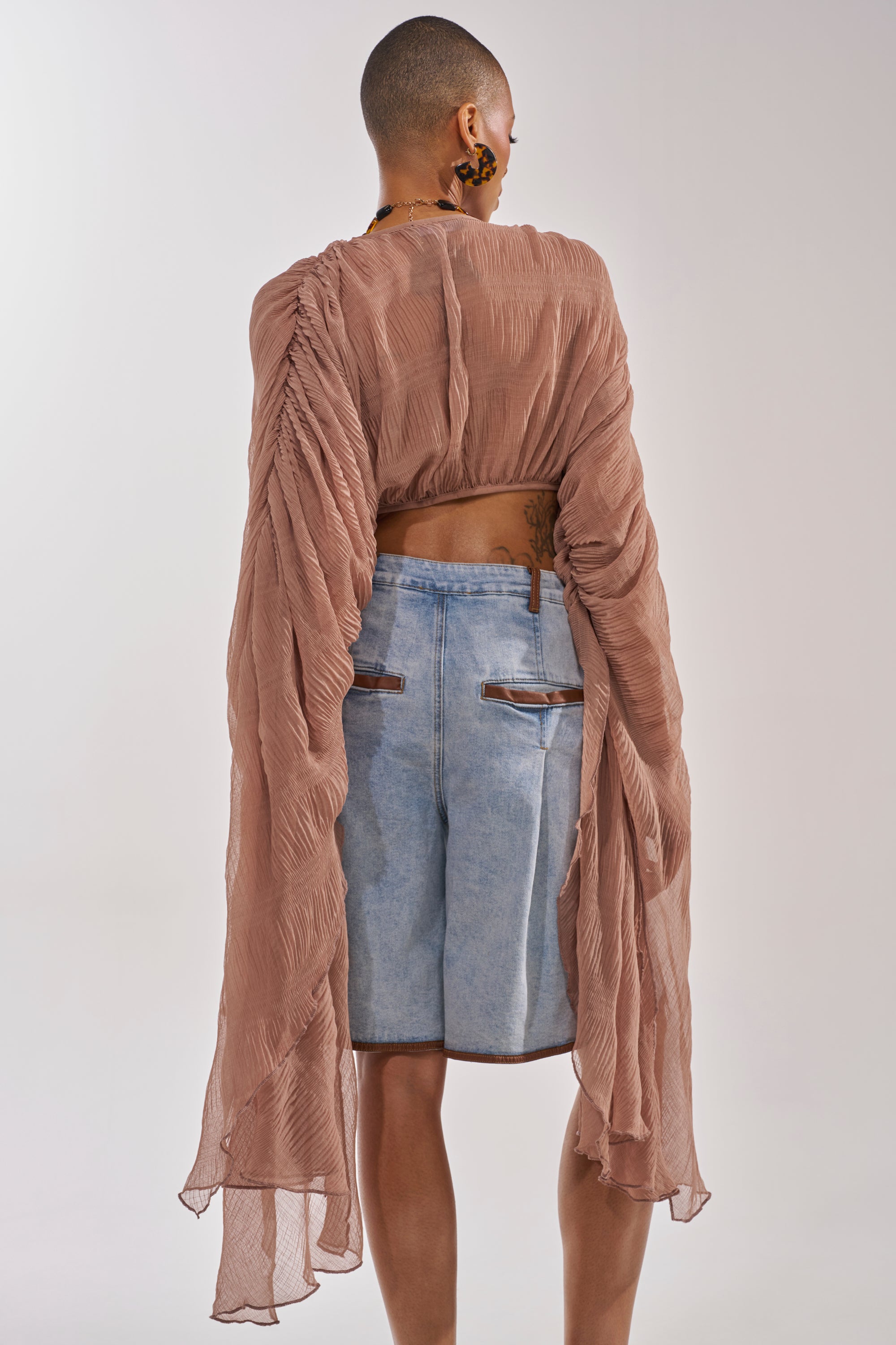 A person stands facing away, wearing the FLY AWAY CHIFFON BLOUSE IN TAUPE with sheer pleated chiffon and draped sleeves, paired with light blue knee-length denim shorts, against a plain light background. 3