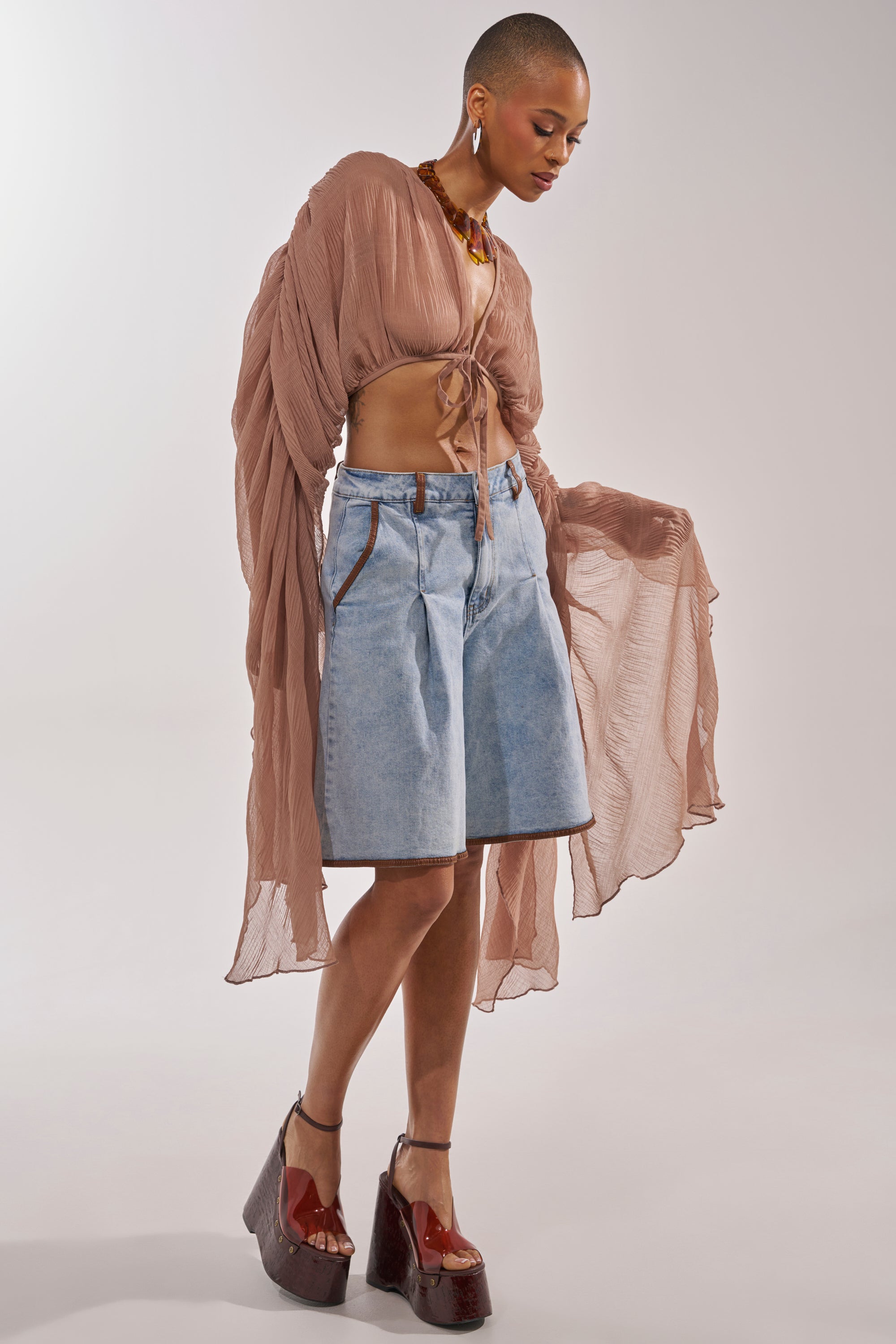 A person poses against a plain background wearing the FLY AWAY CHIFFON BLOUSE IN TAUPE with long sleeves and a tie-front, styled with light blue knee-length denim shorts, chunky brown platform shoes, and a statement necklace. 4