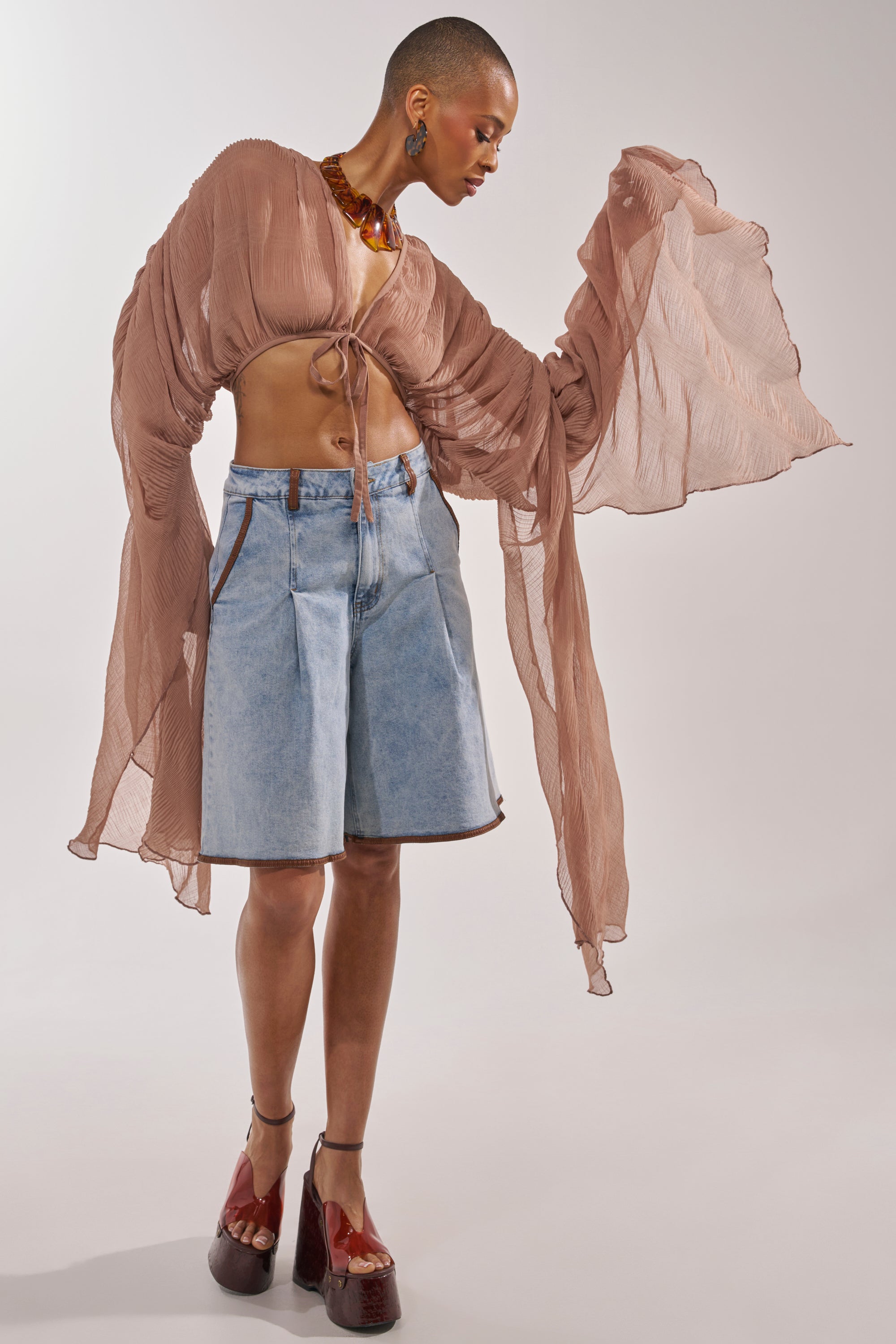 A person with a shaved head models the FLY AWAY CHIFFON BLOUSE IN TAUPE paired with denim knee-length shorts, platform shoes, and a chunky necklace, posing against a plain background. 5