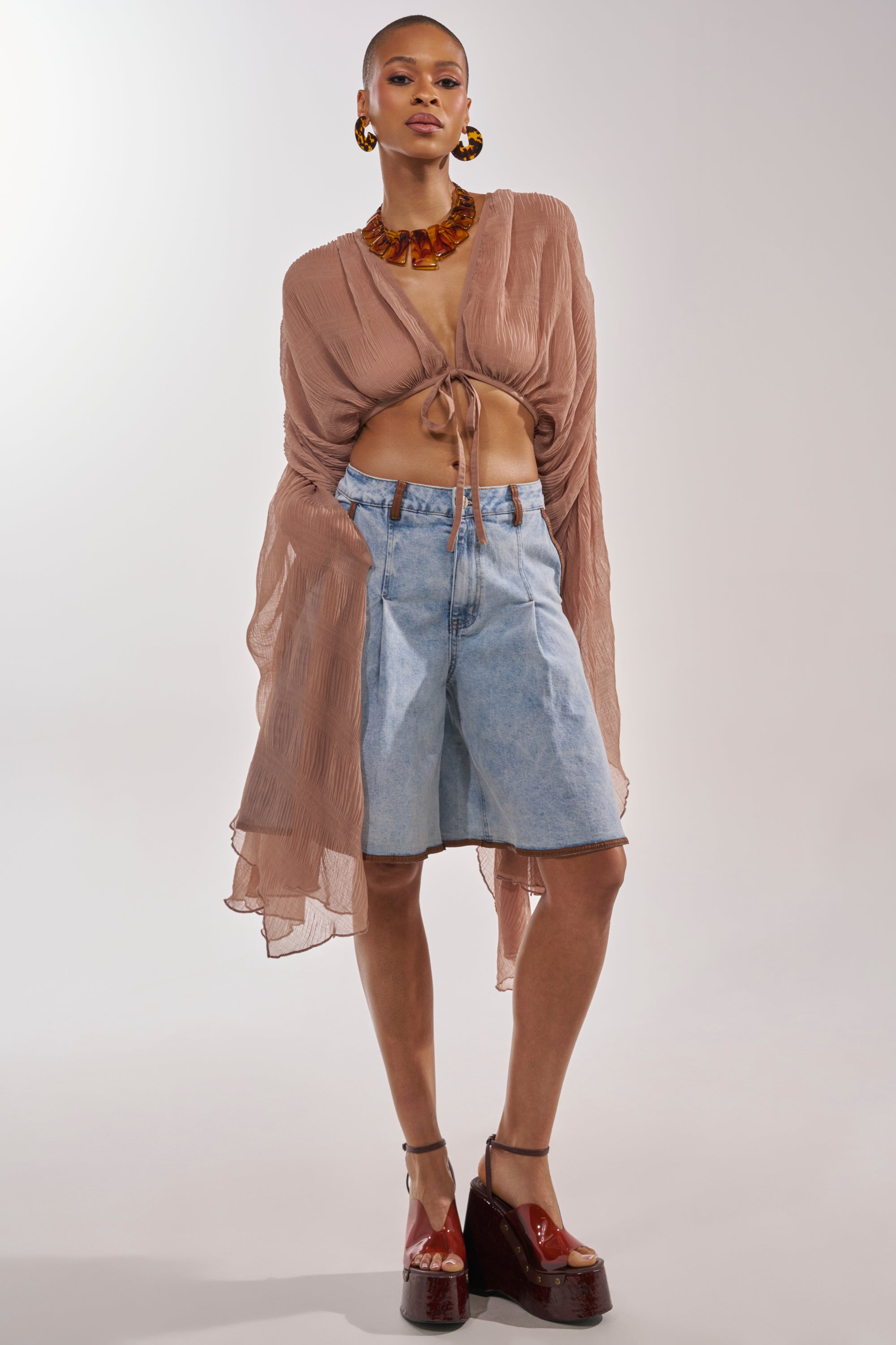 A person stands against a plain background wearing the FLY AWAY CHIFFON BLOUSE IN TAUPE, high-waisted knee-length denim shorts, chunky brown platform shoes, large hoop earrings, and a bold amber necklace. 6