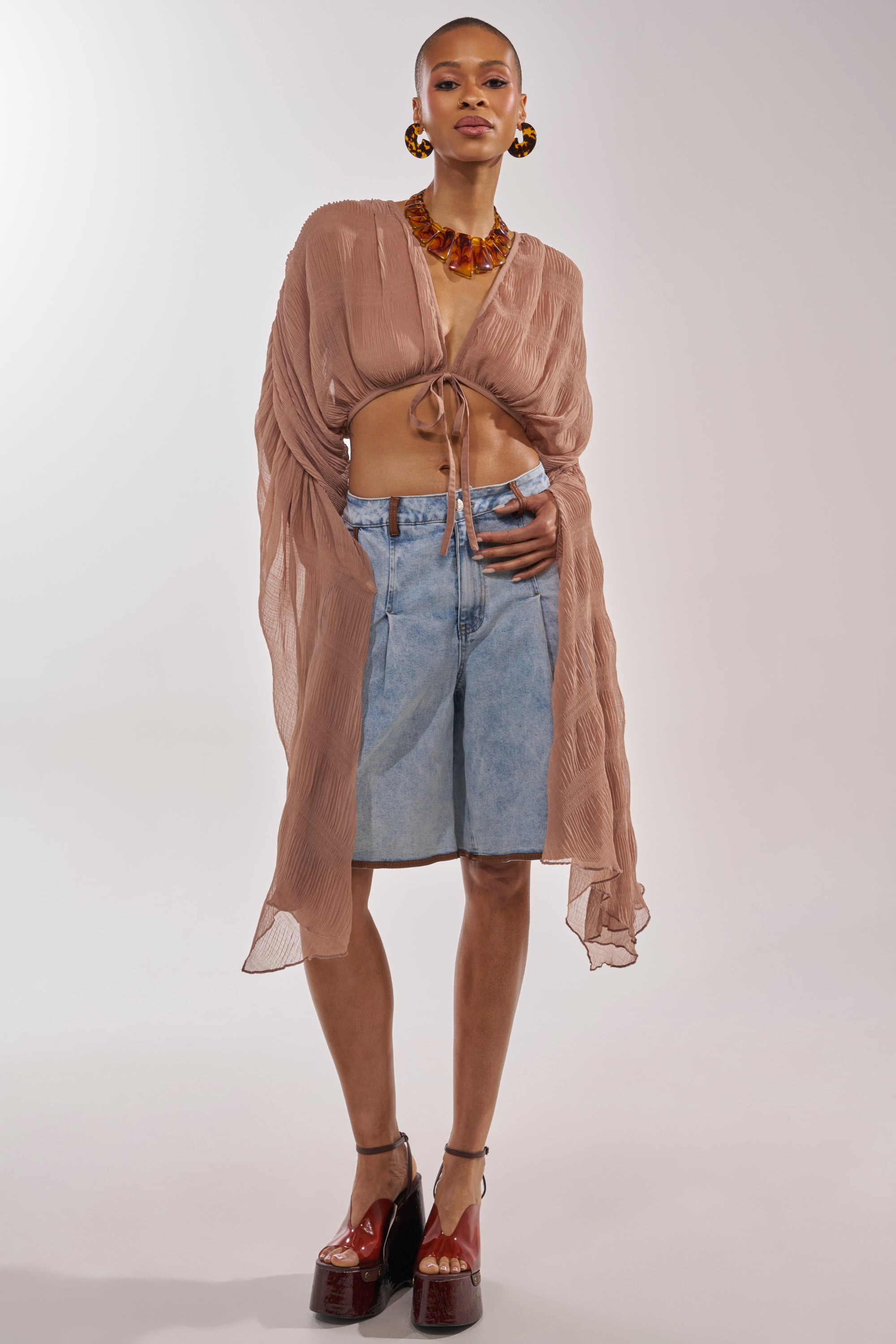 A woman stands against a plain light background wearing the FLY AWAY CHIFFON BLOUSE IN TAUPE, long denim shorts, chunky platform heels, and a large amber-colored necklace with matching earrings. 7