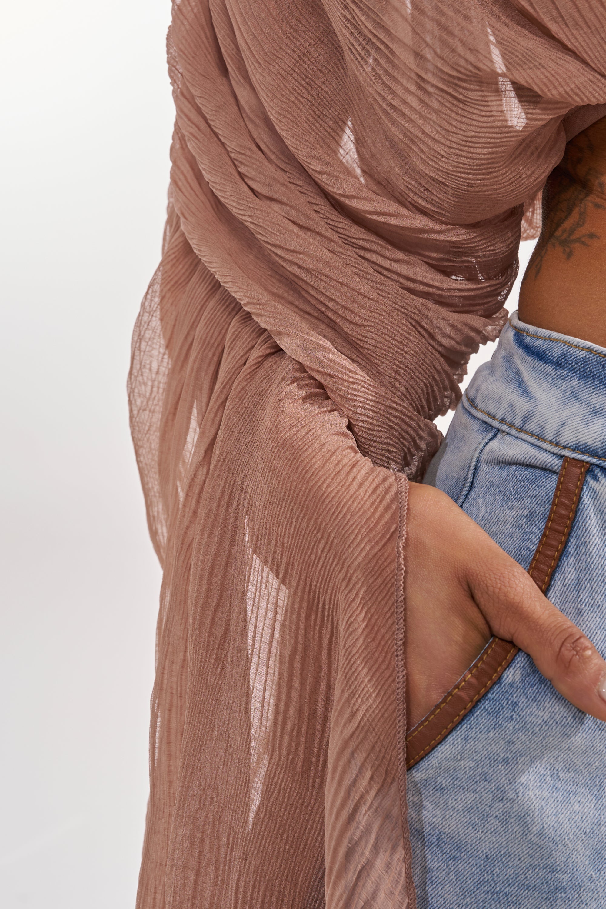 A person in light blue jeans with one hand in their pocket, wearing the FLY AWAY CHIFFON BLOUSE IN TAUPE draped elegantly over their arm and shoulder. 8