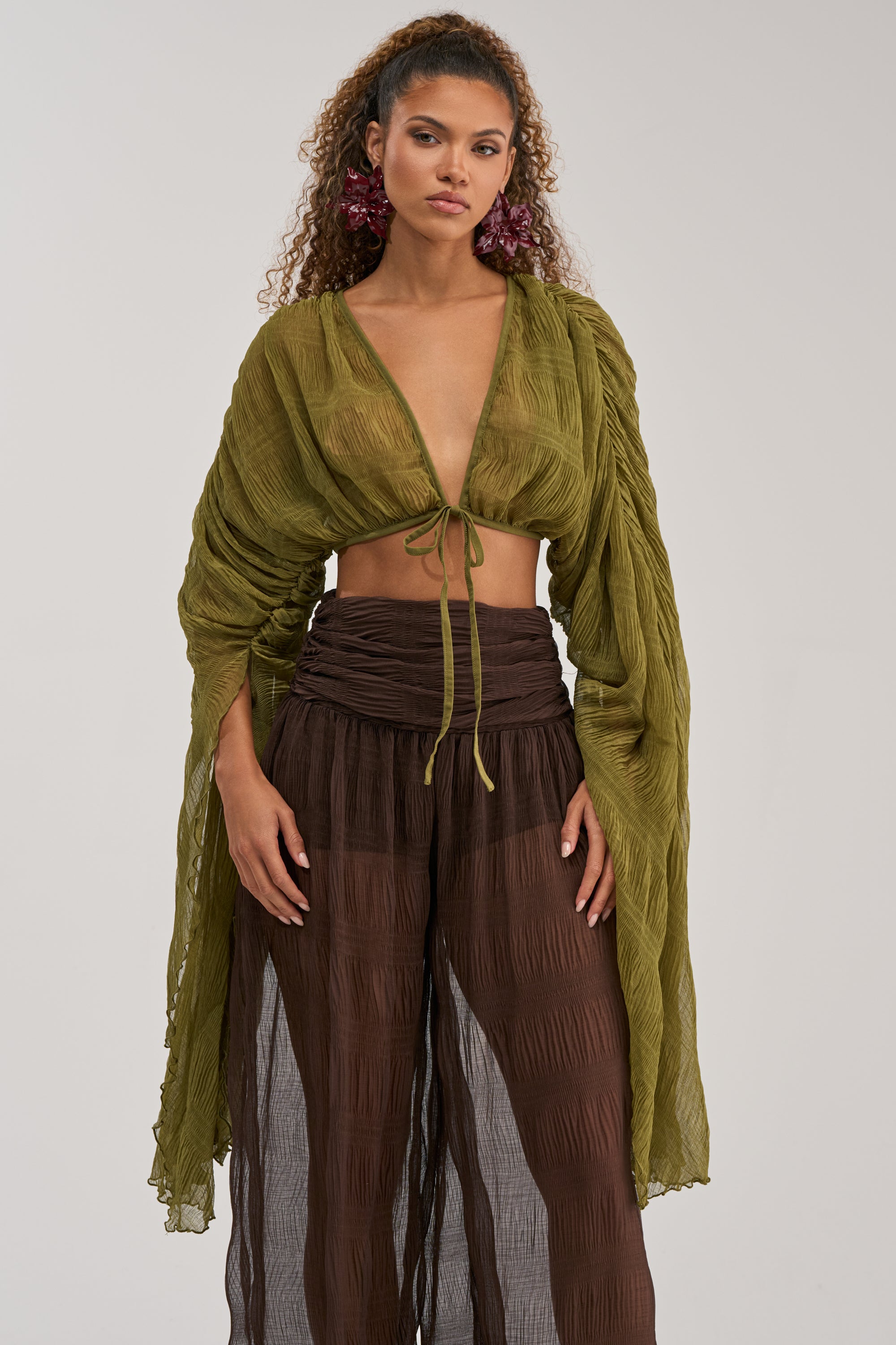 A woman with curly hair and large pink earrings stands against a plain background, wearing the FORBIDDEN FOREST CHIFFON BLOUSE—a sheer olive green pleated chiffon top with long sleeves and a tie front—paired with high-waisted, semi-transparent brown pants.