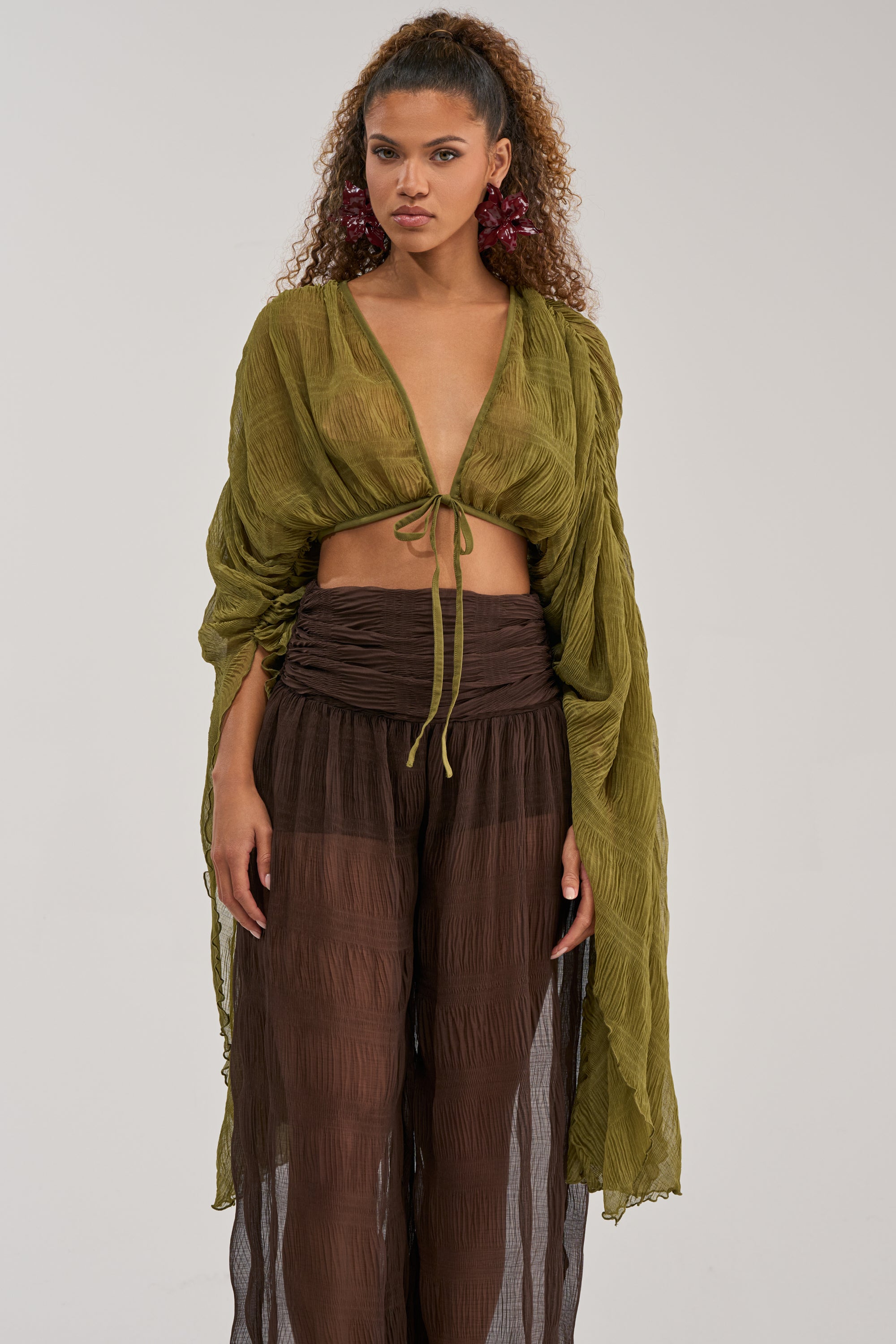 A woman wearing the FORBIDDEN FOREST CHIFFON BLOUSE—an olive cropped blouse featuring sheer long sleeves and a deep V-neck tie front—paired with brown sheer pants, stands against a plain background.