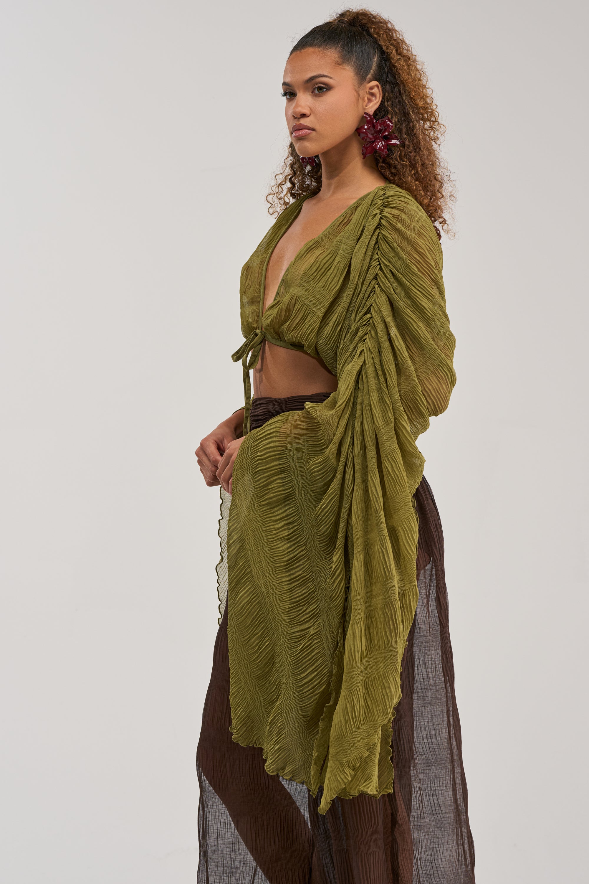 A woman with curly hair in a ponytail wears the FORBIDDEN FOREST CHIFFON BLOUSE, styled as an olive green textured wrap top with long, ruched sleeves, paired with brown sheer wide-leg pants, standing against a plain background.