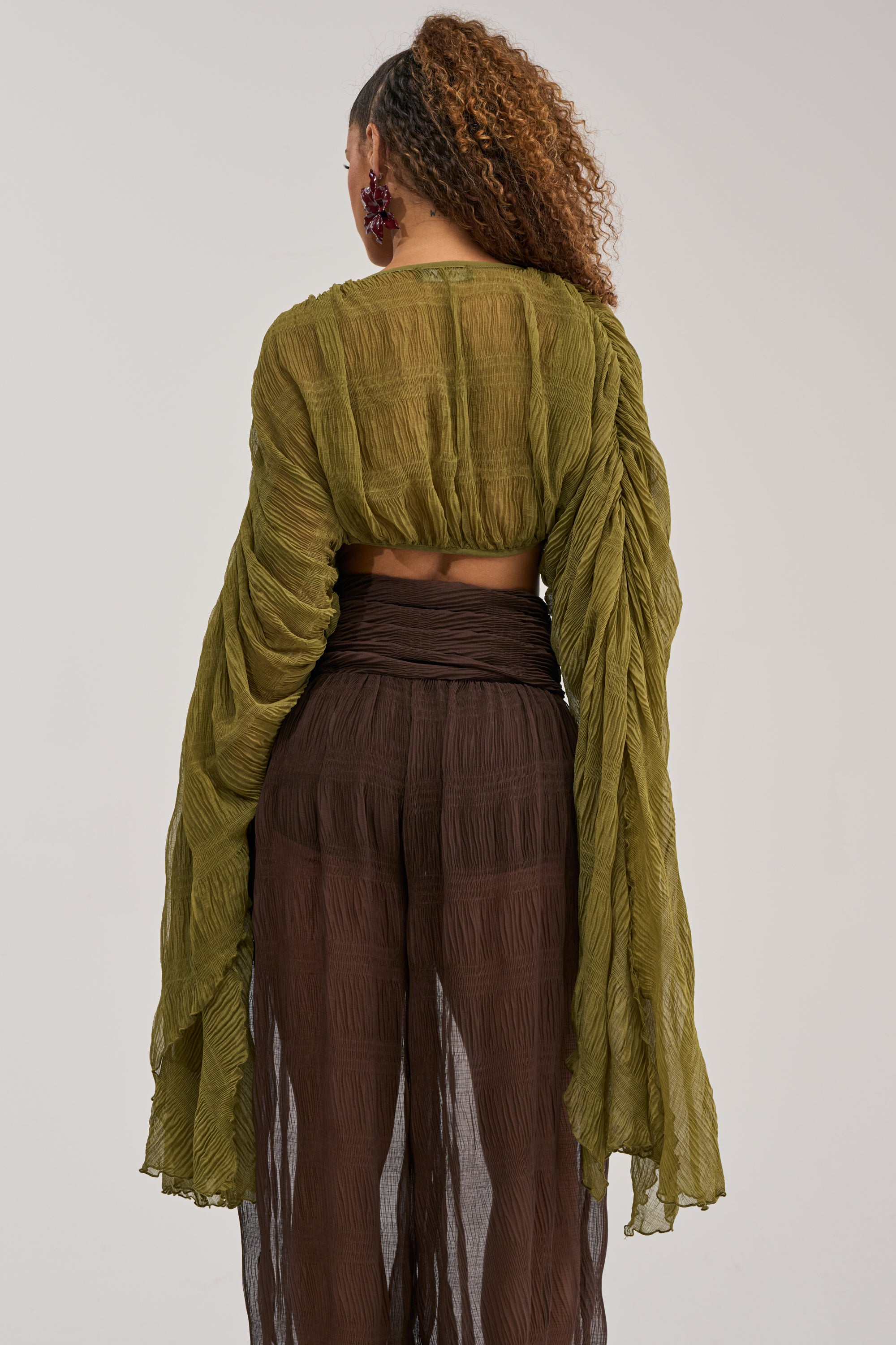 A person with curly hair wears the FORBIDDEN FOREST CHIFFON BLOUSE in olive, featuring long sleeves, paired with dark brown sheer pants. They are standing with their back to the camera against a plain background.