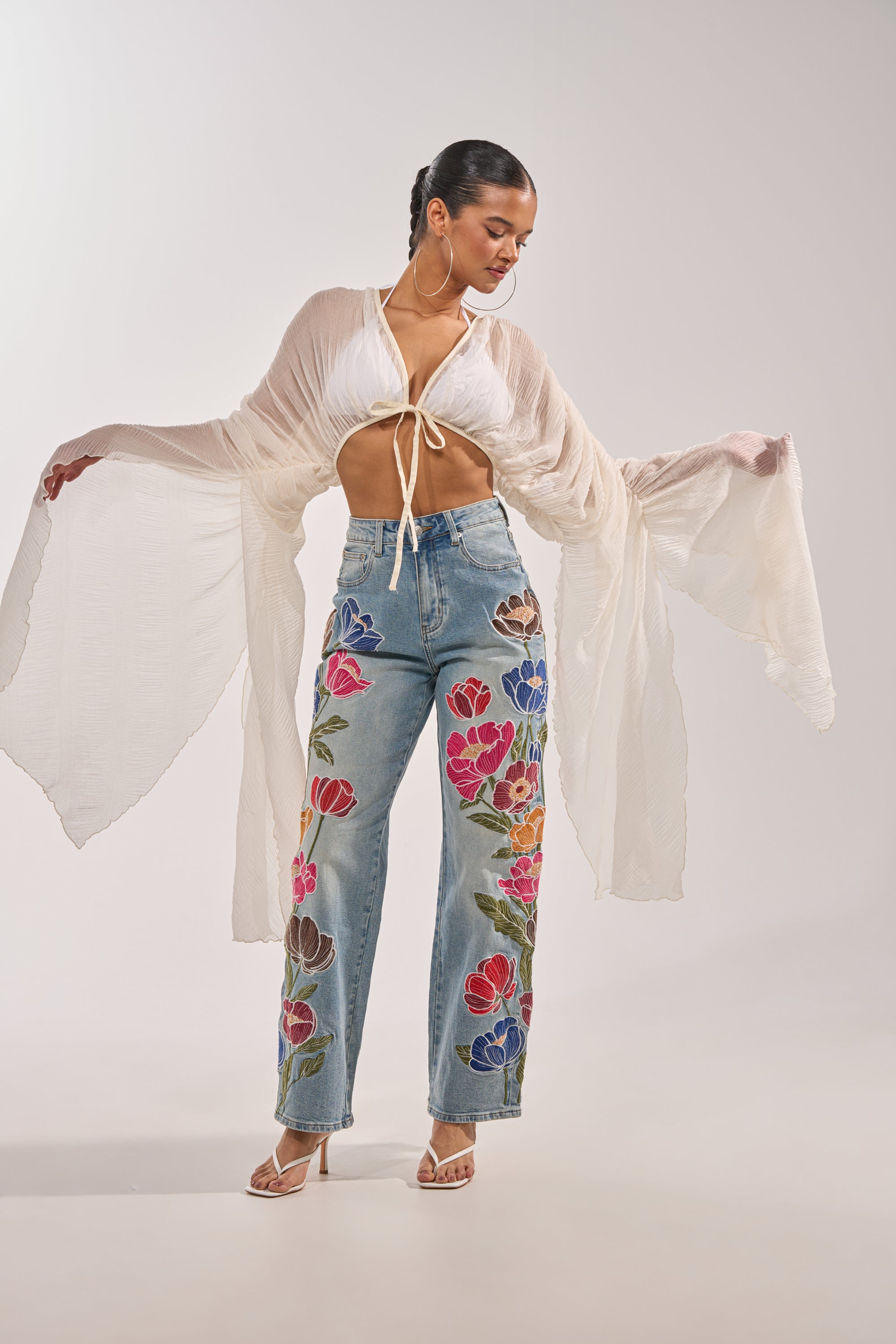 A woman models the FLY AWAY CHIFFON BLOUSE—a sheer, pleated top with wide sleeves—styled with a white halter bralette, embroidered high-waisted jeans, heeled sandals, and her hair pulled back. 0
