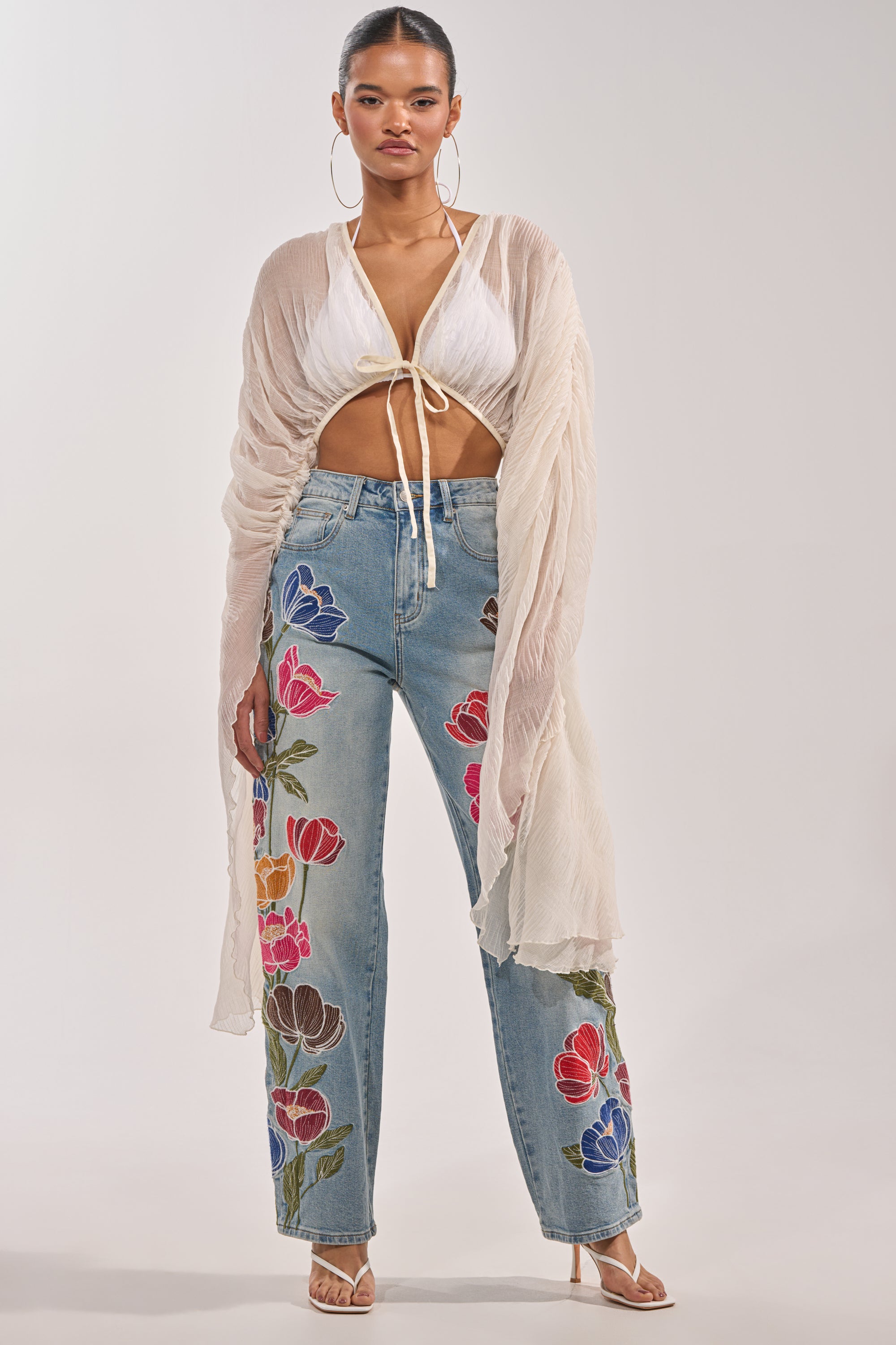 A woman wears the FLY AWAY CHIFFON BLOUSE—a sheer ivory pleated top with a deep V-neckline—over a white crop top and embroidered high-waisted jeans, styled with strappy heels and hoop earrings for a chic festival look. 1