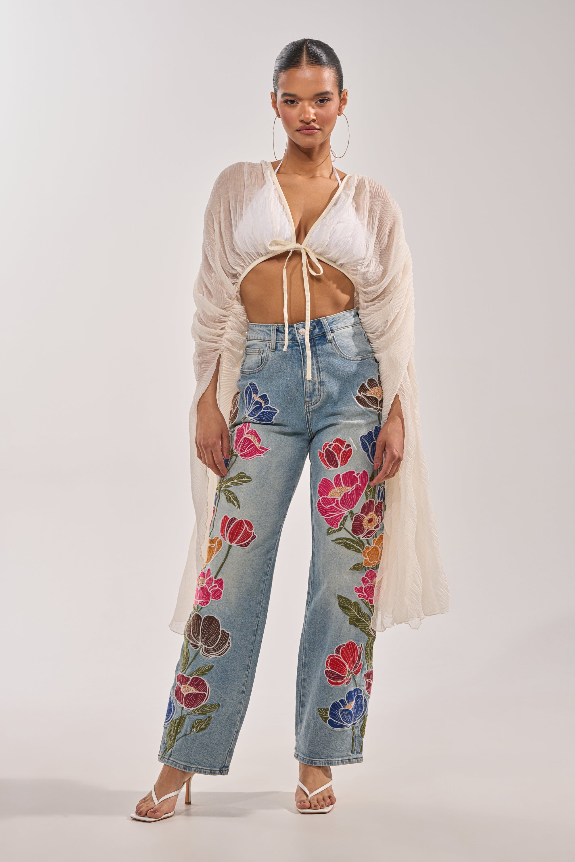 A woman poses in the FLY AWAY CHIFFON BLOUSE—a sheer ivory pleated top layered over a white halter—paired with light blue embroidered jeans and white high-heeled sandals against a plain background. 2
