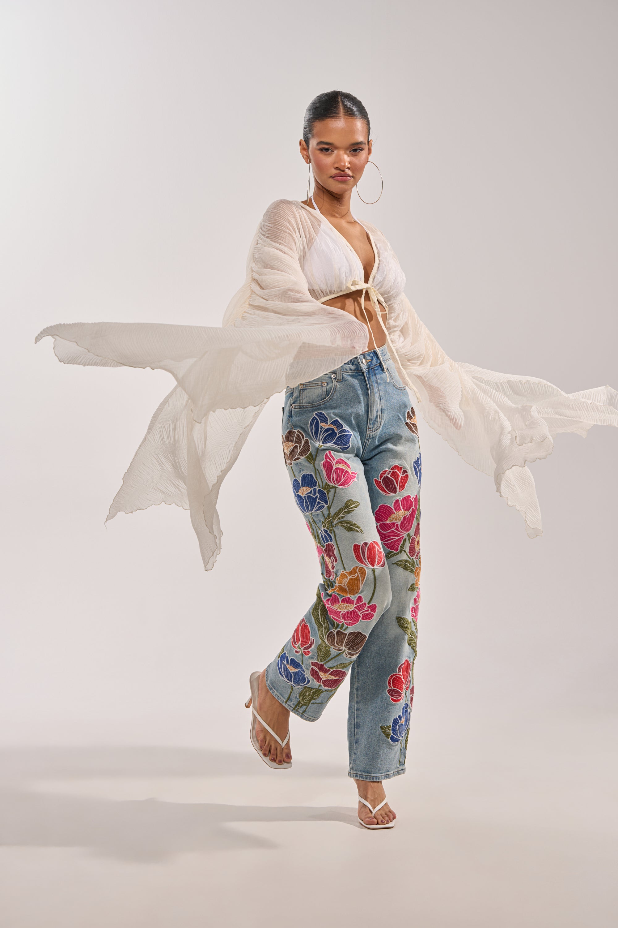 A woman models the FLY AWAY CHIFFON BLOUSE, a sheer ivory pleated top with a deep V-neckline, paired with blue floral embroidered jeans, hoop earrings, and heeled sandals against a plain light background. 3