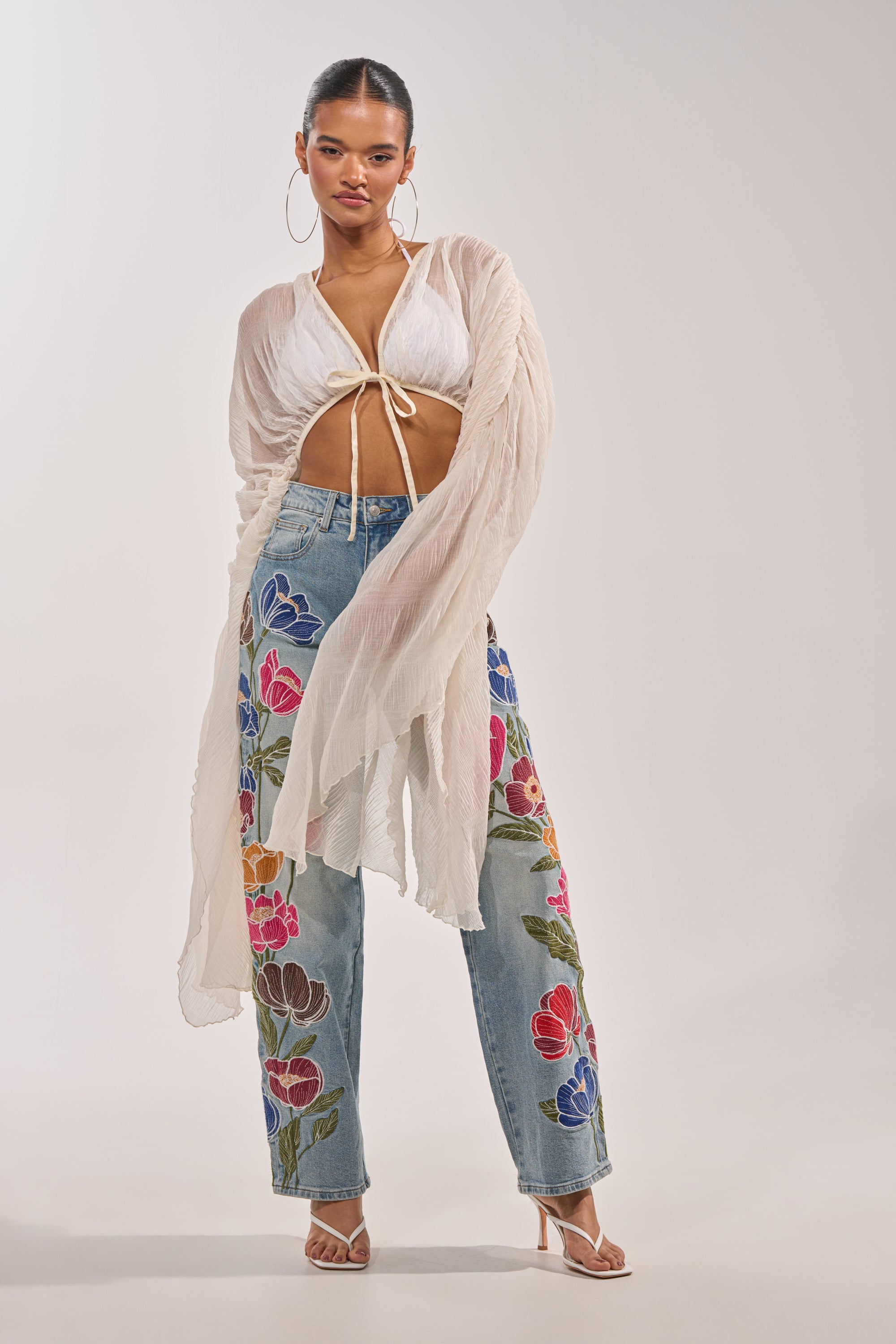 A woman poses in the FLY AWAY CHIFFON BLOUSE—an ivory sheer pleated top with a deep V-neckline, styled over a white bralette and high-waisted embroidered jeans, making for an ideal festival look with strappy heeled sandals. 4
