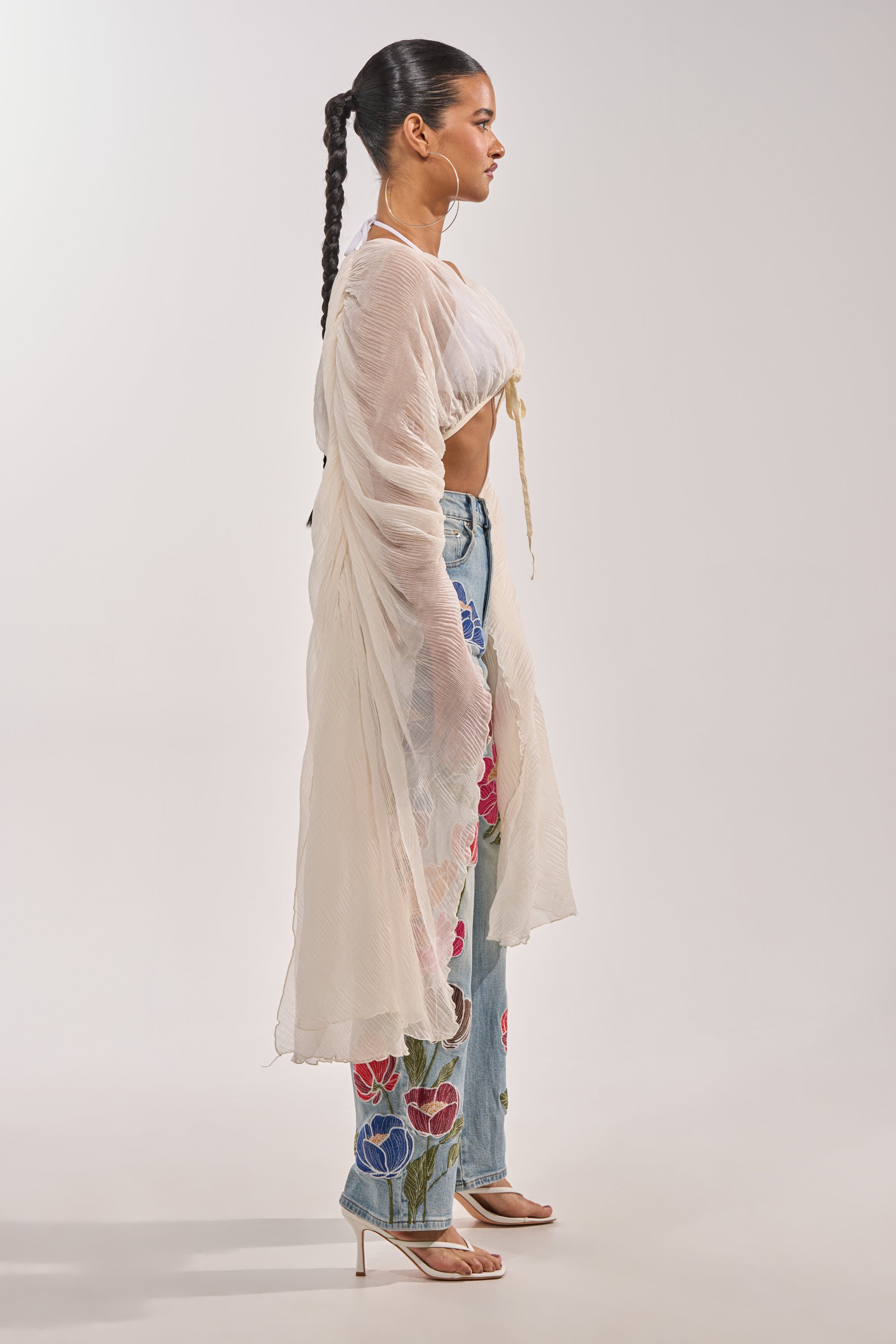 A woman stands in profile, wearing the FLY AWAY CHIFFON BLOUSE with blue embroidered jeans and beige heels—a stylish, breezy festival look against a simple background. 5