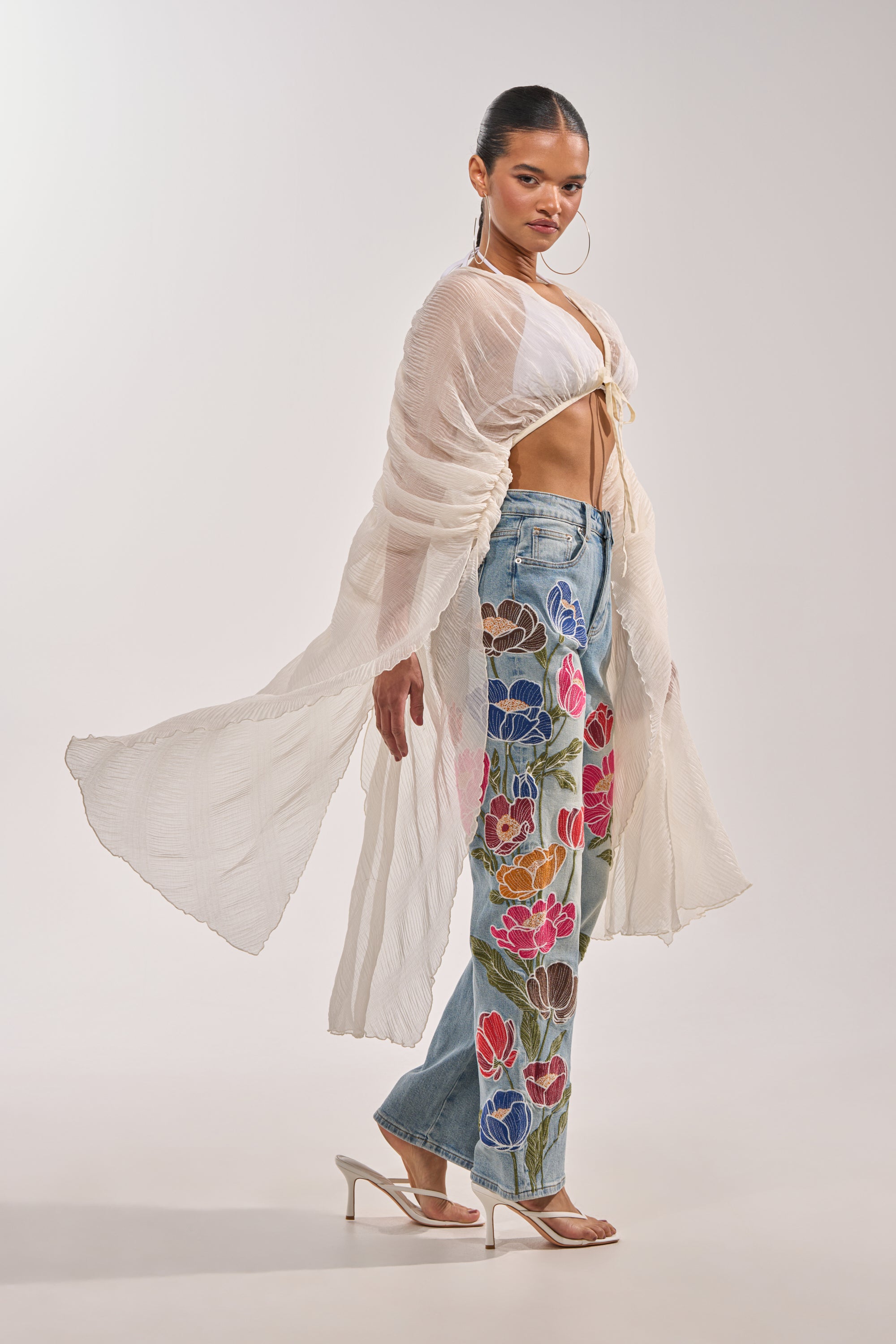 A woman poses against a plain light background in the FLY AWAY CHIFFON BLOUSE—a sheer pleated top with a deep V-neck—paired with floral blue jeans, clear heels, large hoop earrings, and pulled-back hair for a chic festival look. 6