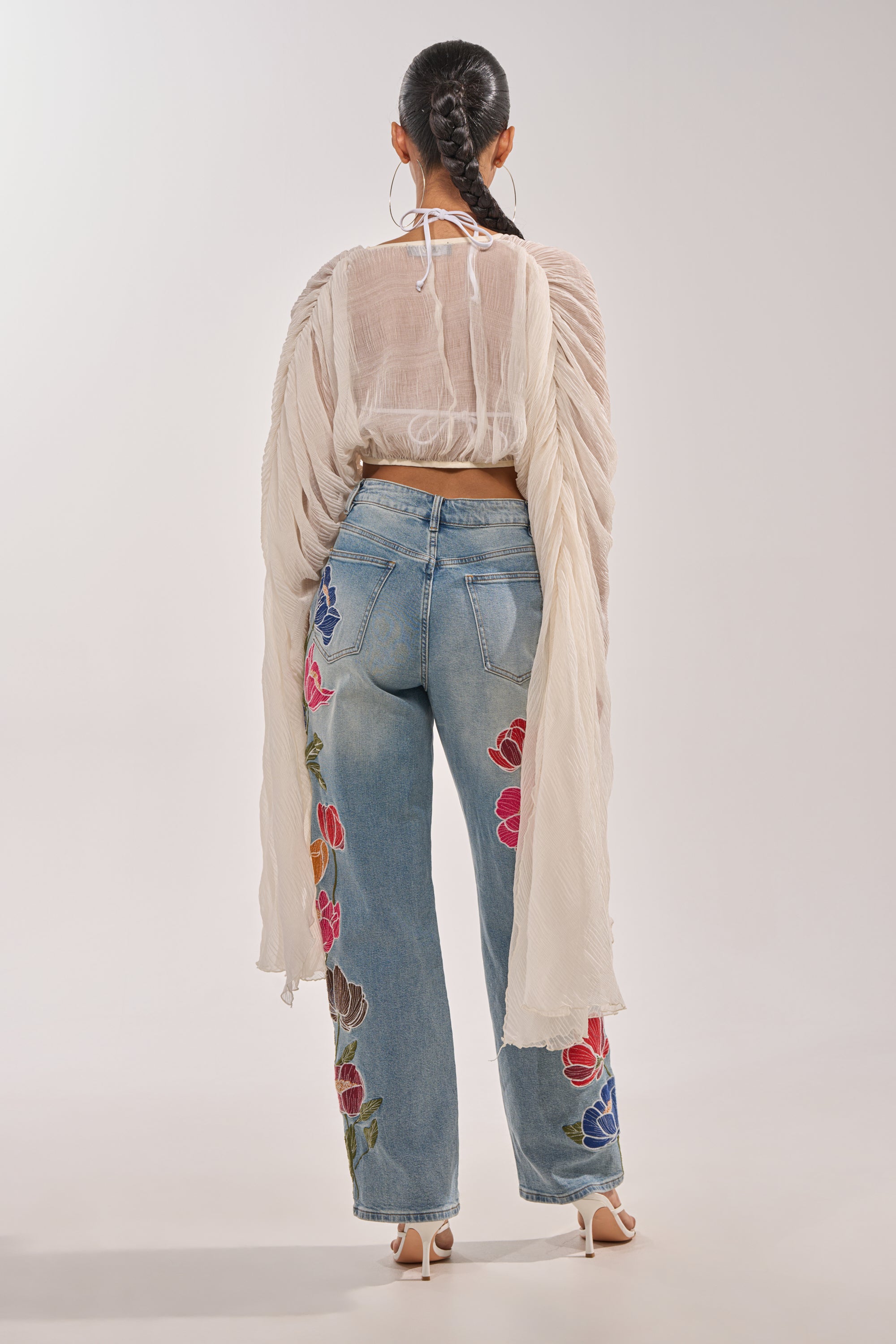 A woman with braided hair faces away, showcasing a festival look in the FLY AWAY CHIFFON BLOUSE with dramatic draped sleeves, paired with light blue high-waisted jeans featuring flower embroidery and finished with white heels. 7