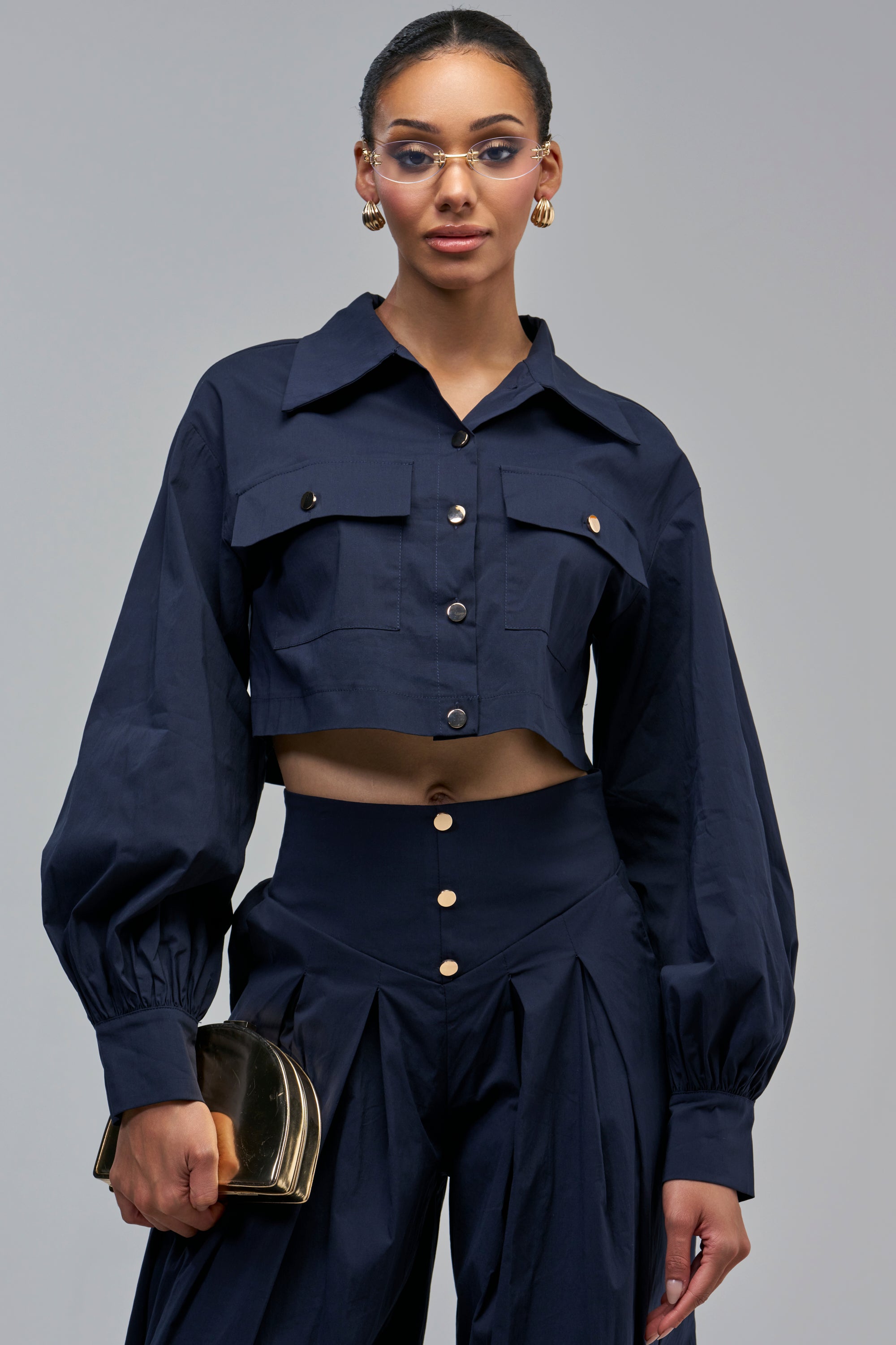 A person wearing a navy ALL ABOUT ME BUTTON DOWN with a cropped silhouette and voluminous balloon sleeves paired with matching high-waisted trousers, holding a gold clutch.