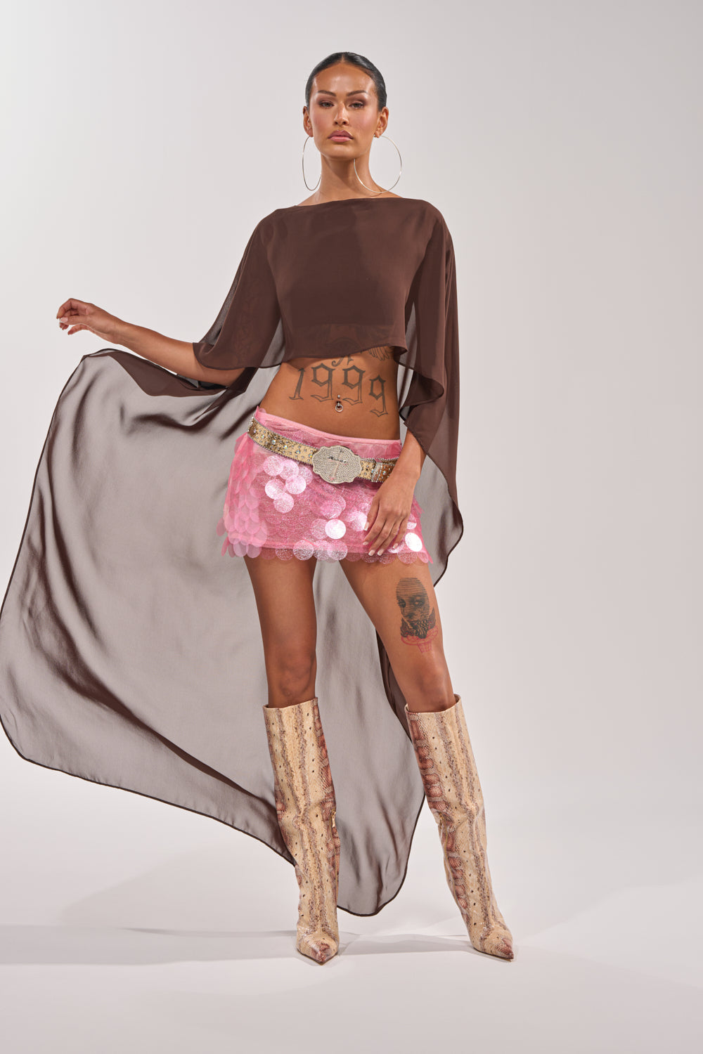 A woman poses confidently in the IT GIRL BLOUSE IN BROWN, styled with a pink sequin mini skirt, wide belt, tall patterned boots, and large hoop earrings. Her midriff and thigh tattoos are visible under the elegant evening resort look. 2