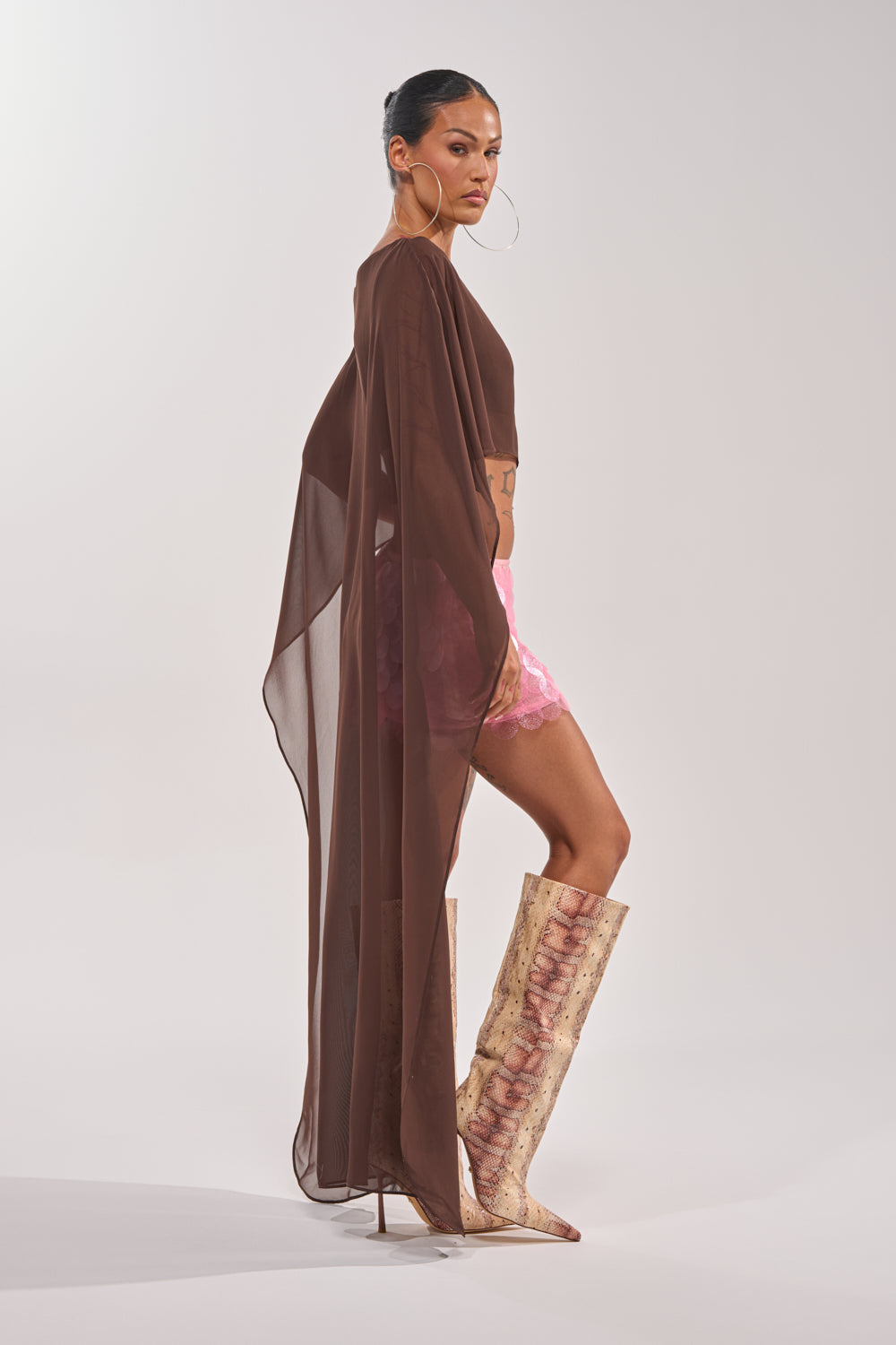 A woman models the IT GIRL BLOUSE IN BROWN, a sheer chiffon top with a high-low hem, paired with pink lace shorts, hoop earrings, and tall textured boots. She stands in profile against a plain light background. 5