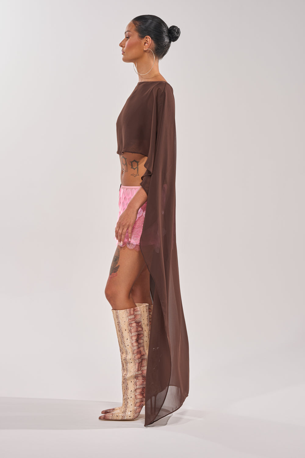 A person stands in profile against a plain background, wearing the IT GIRL BLOUSE IN BROWN, pink shorts, tall beige textured boots, and showcasing visible tattoos. 6