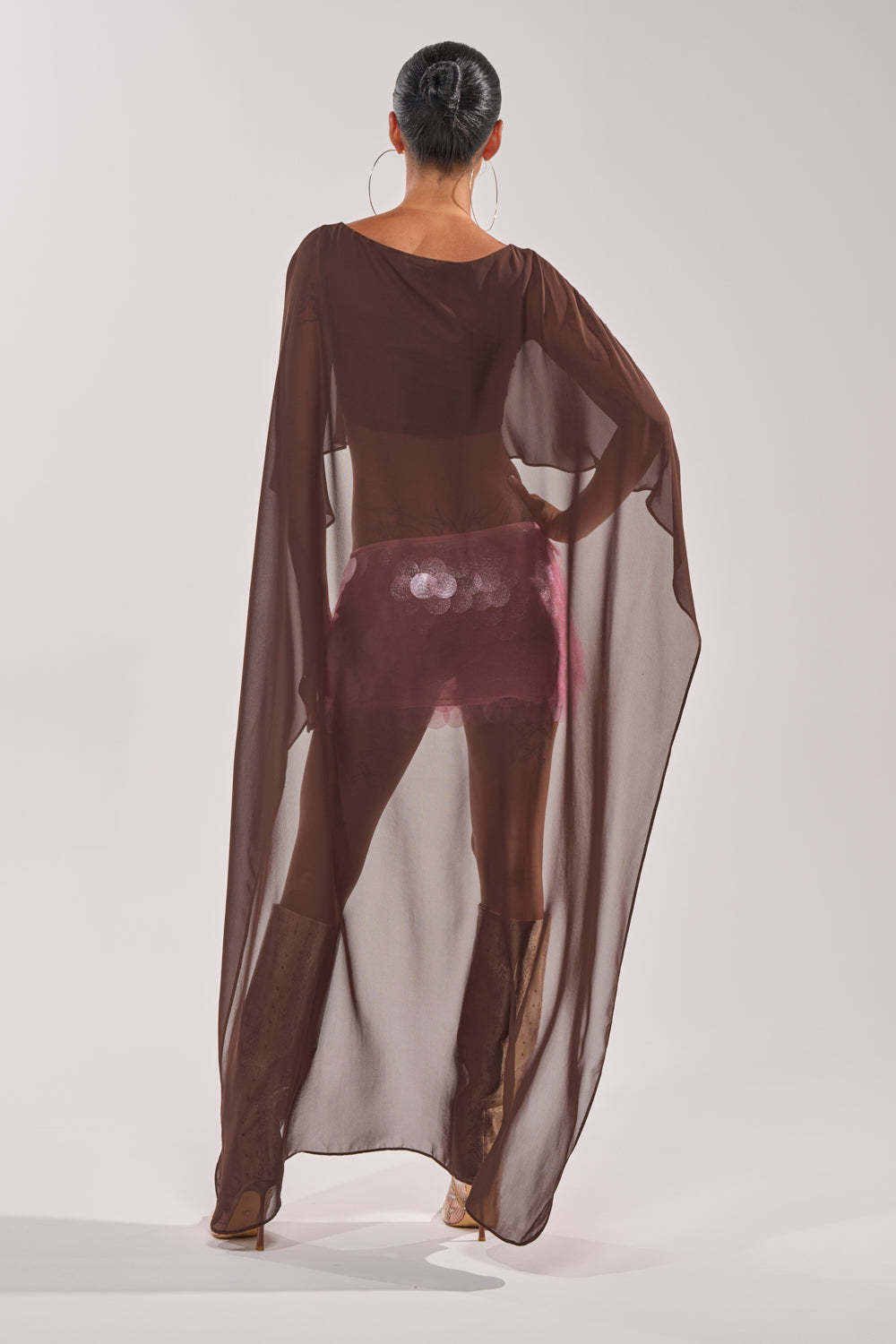A person stands facing away, wearing the IT GIRL BLOUSE IN BROWN—a sheer chiffon top with wide sleeves and high-low hem—over shiny pink shorts. Large hoop earrings, brown boots, and a neat bun finish this perfect resort-wear look. 7
