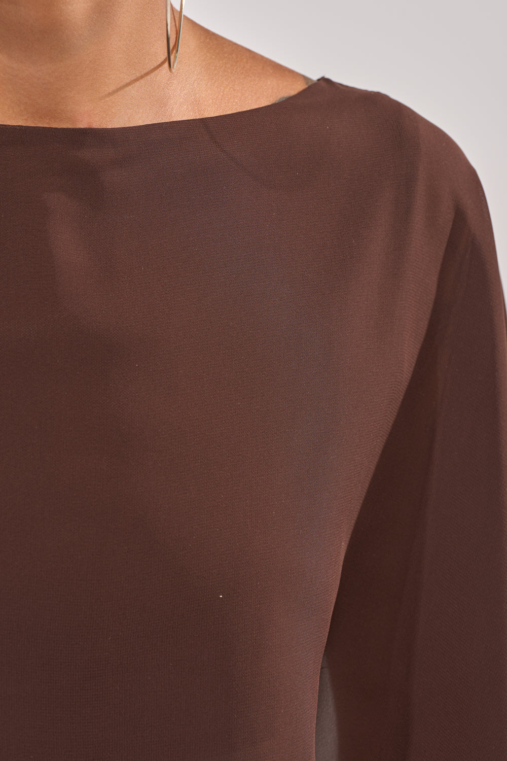 Close-up of a person wearing the IT GIRL BLOUSE IN BROWN, a sheer chiffon top with long sleeves, wide neckline, high-low hem, and paired with a thin metallic hoop earring—ideal for resort wear. Only the upper chest, shoulder, and neck are shown. 8