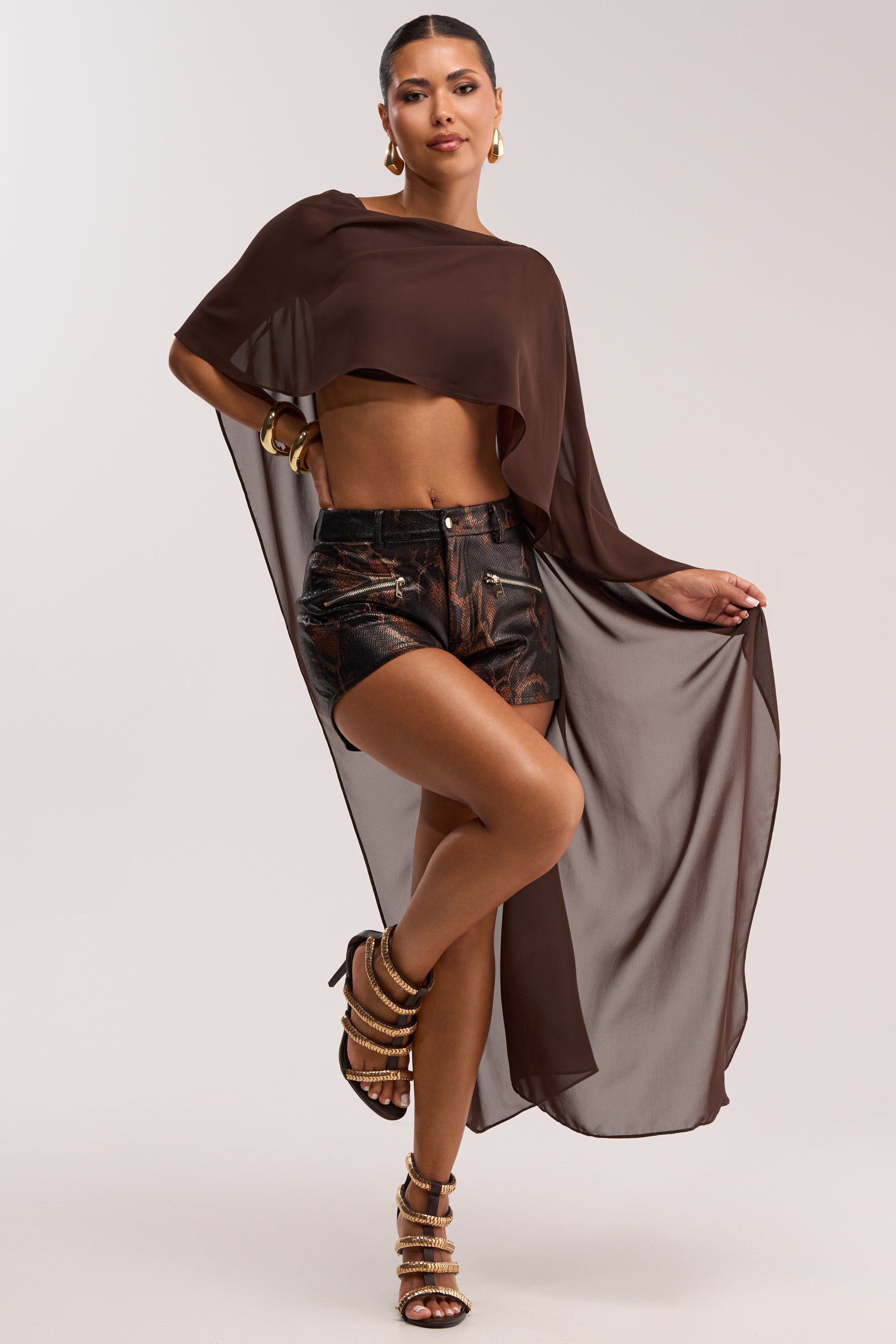 A woman poses in chic evening resort wear, featuring the IT GIRL BLOUSE IN BROWN— a sheer brown chiffon top with an asymmetrical high-low hem, styled with metallic shorts, strappy heels, and bold gold accessories.