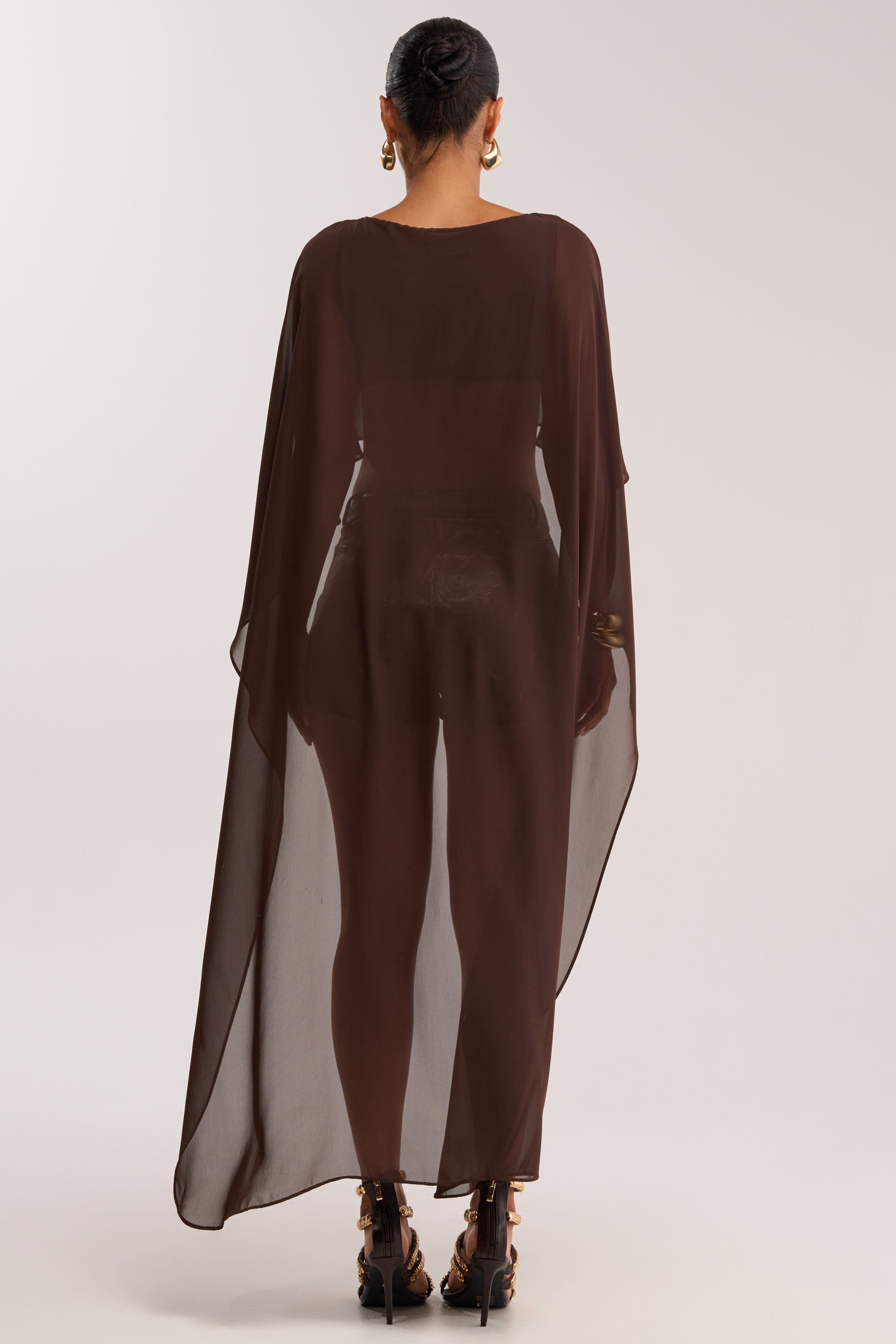 A person stands facing away, wearing the IT GIRL BLOUSE IN BROWN—a sheer chiffon top with a high-low hem over black attire, styled with a bun, large hoop earrings, and strappy heels for chic evening resort wear.
