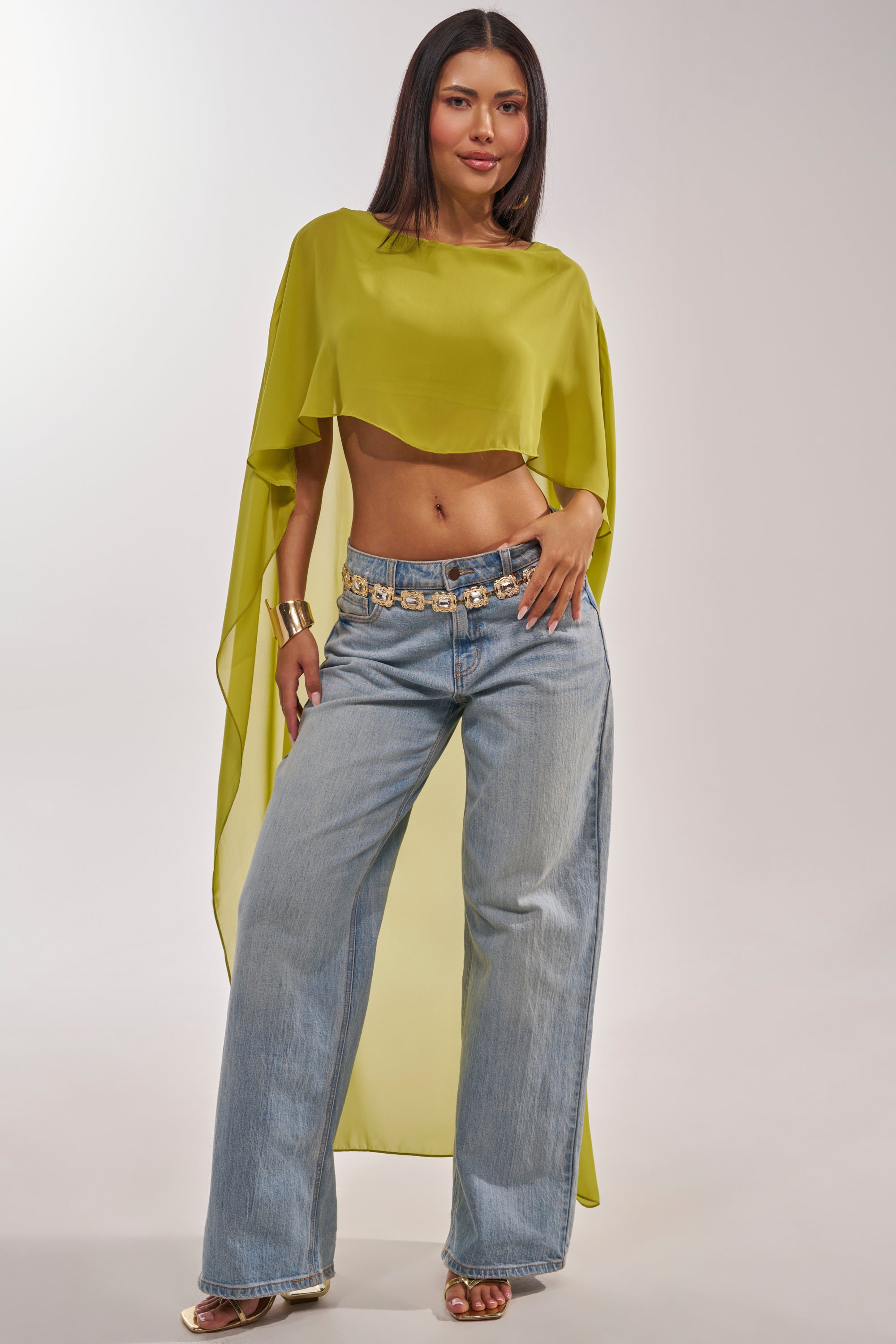 A woman poses confidently in the IT GIRL BLOUSE IN CHARTREUSE and wide-leg jeans, styled with a gold belt and bracelet, against a light neutral background for fresh resort wear inspiration. 1