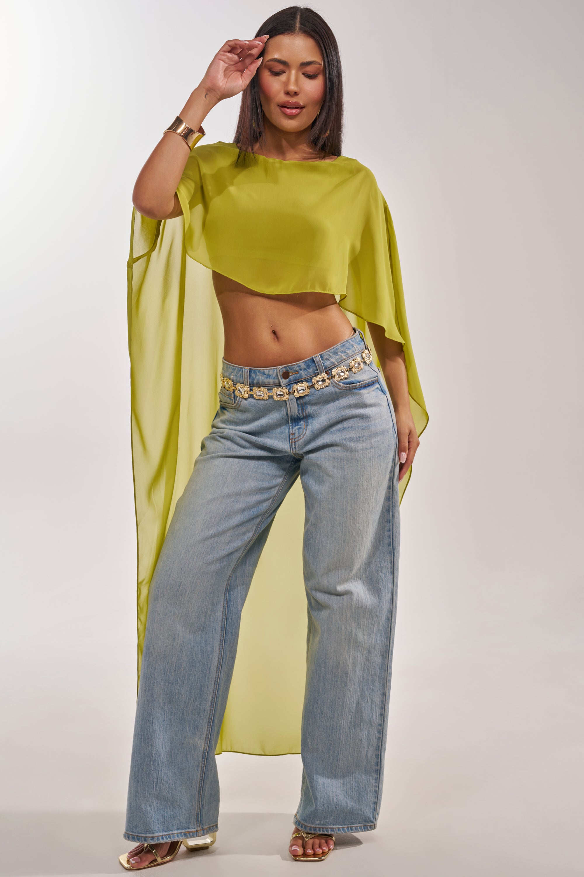 A woman models the IT GIRL BLOUSE IN CHARTREUSE—a sheer chiffon top with flowy sleeves—styled with wide-leg light blue jeans, gold sandals, a statement gold belt, and matching bracelets for chic resort wear against a plain background. 2
