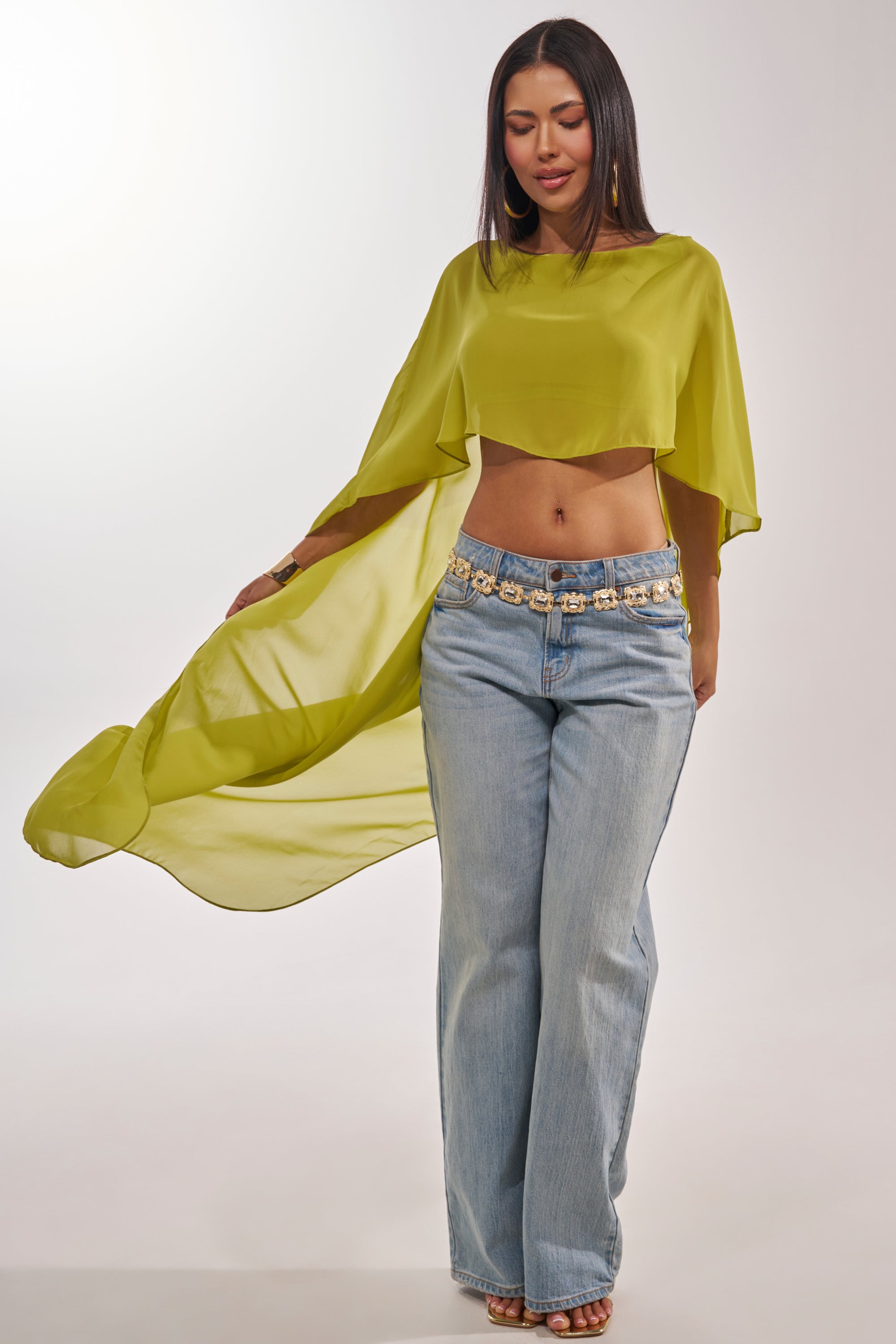 A woman stands confidently in the IT GIRL BLOUSE IN CHARTREUSE, a flowing chiffon top styled for resort wear, paired with light blue wide-leg jeans. She holds out the vibrant high-low blouse, her straight hair and decorated belt completing the chic look. 3