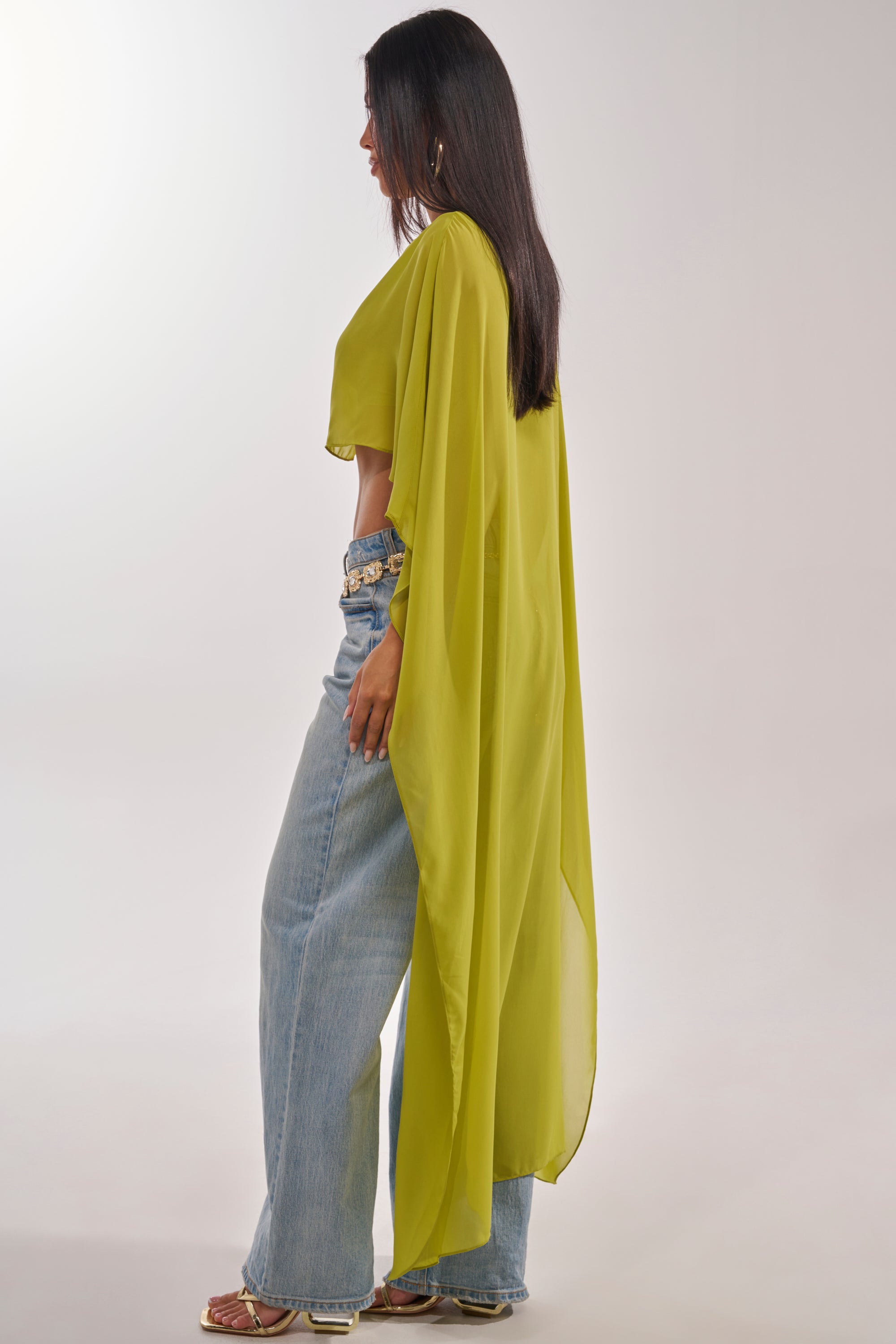 A woman stands sideways in the IT GIRL BLOUSE IN CHARTREUSE, styled with a sheer cape, light blue high-waisted jeans, and sandals. The light background highlights her modern, effortlessly stylish resort look. 4