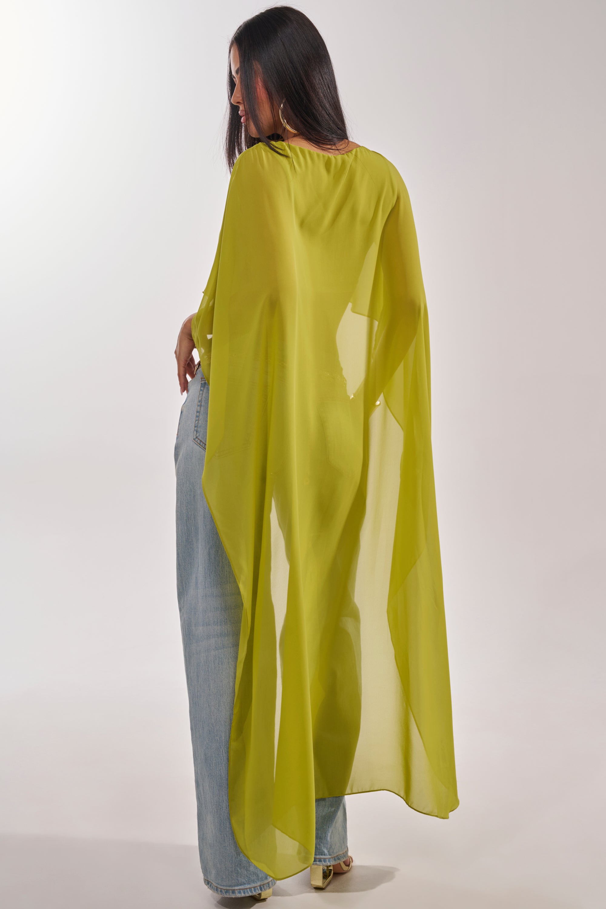 A woman with long dark hair faces away, wearing the IT GIRL BLOUSE IN CHARTREUSE as a sheer cape over light blue wide-leg jeans and heels. She stands against a plain, light background. 5