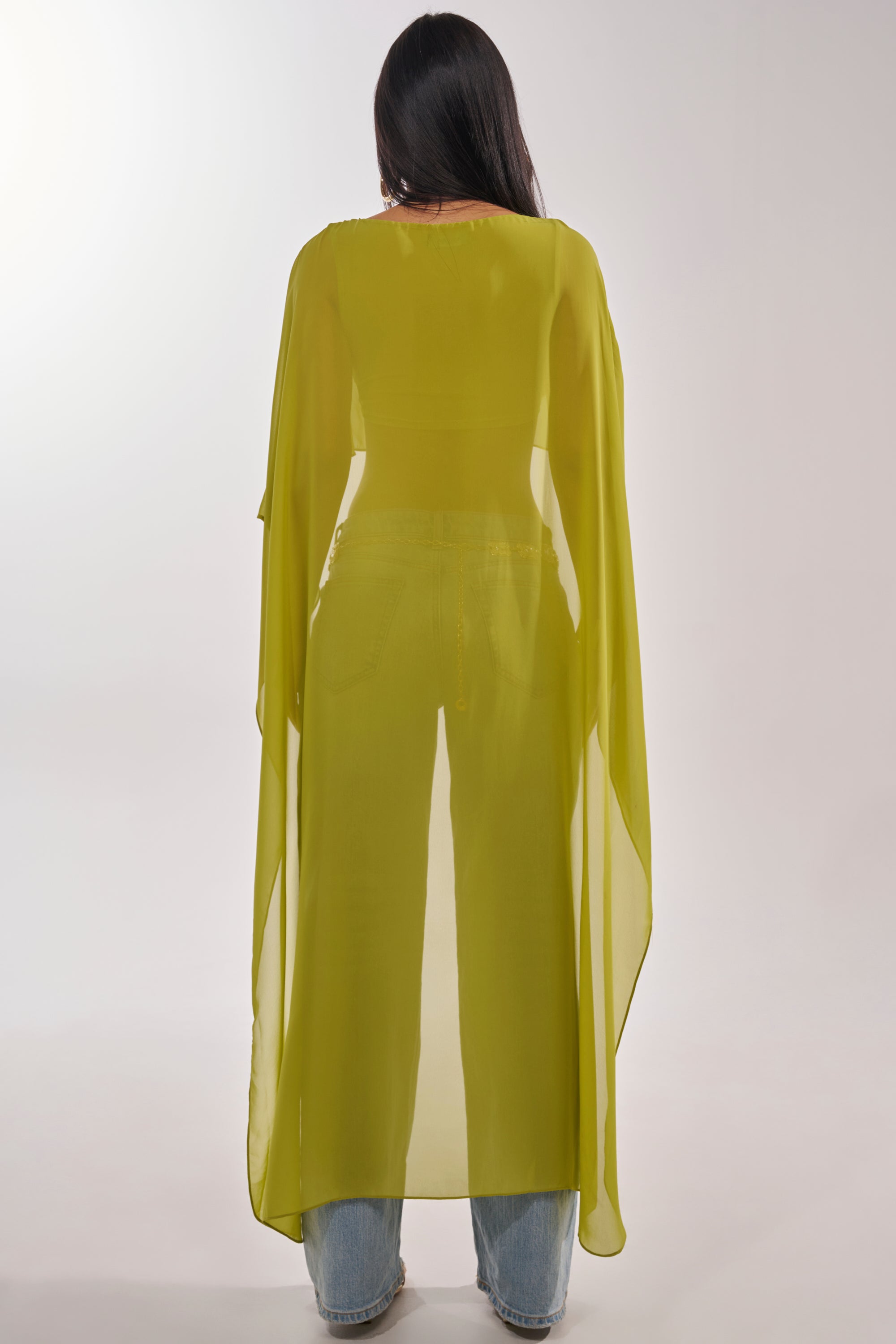 A woman with long dark hair faces away, modeling the IT GIRL BLOUSE IN CHARTREUSE—a sheer chiffon, high-low top with flowy sleeves over blue jeans. The light, ankle-length blouse is perfect for resort wear against a plain background. 6