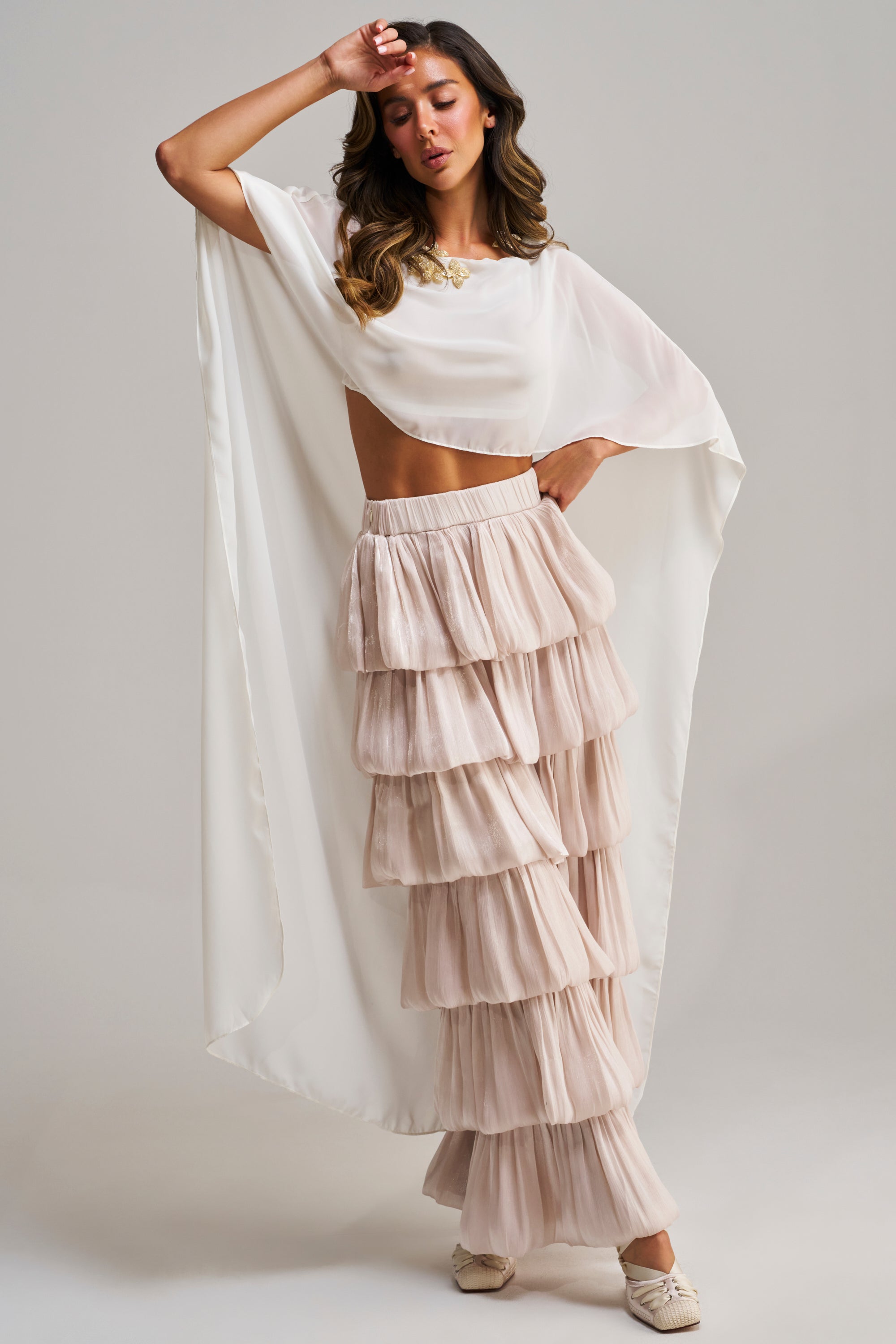 A woman poses in the IT GIRL BLOUSE IN IVORY, a chic chiffon cape top, paired with tiered blush pink ruffle pants. She raises one arm to her forehead and rests the other on her hip against a plain background.