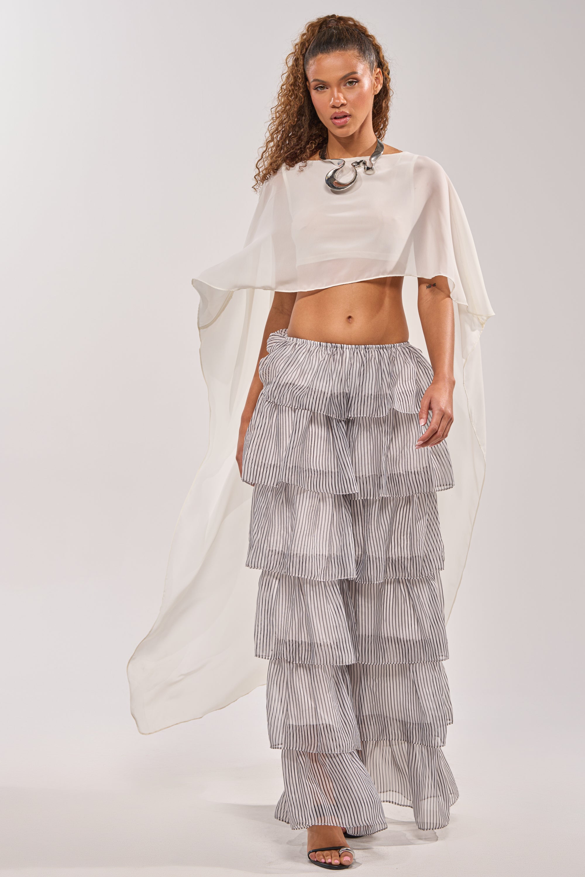 A woman models the IT GIRL BLOUSE IN IVORY, a sheer chiffon cape top with a high-low drape, paired with a ruffled striped skirt, chunky silver necklace, and open-toe heels against a plain backdrop—perfect for festivals. 0