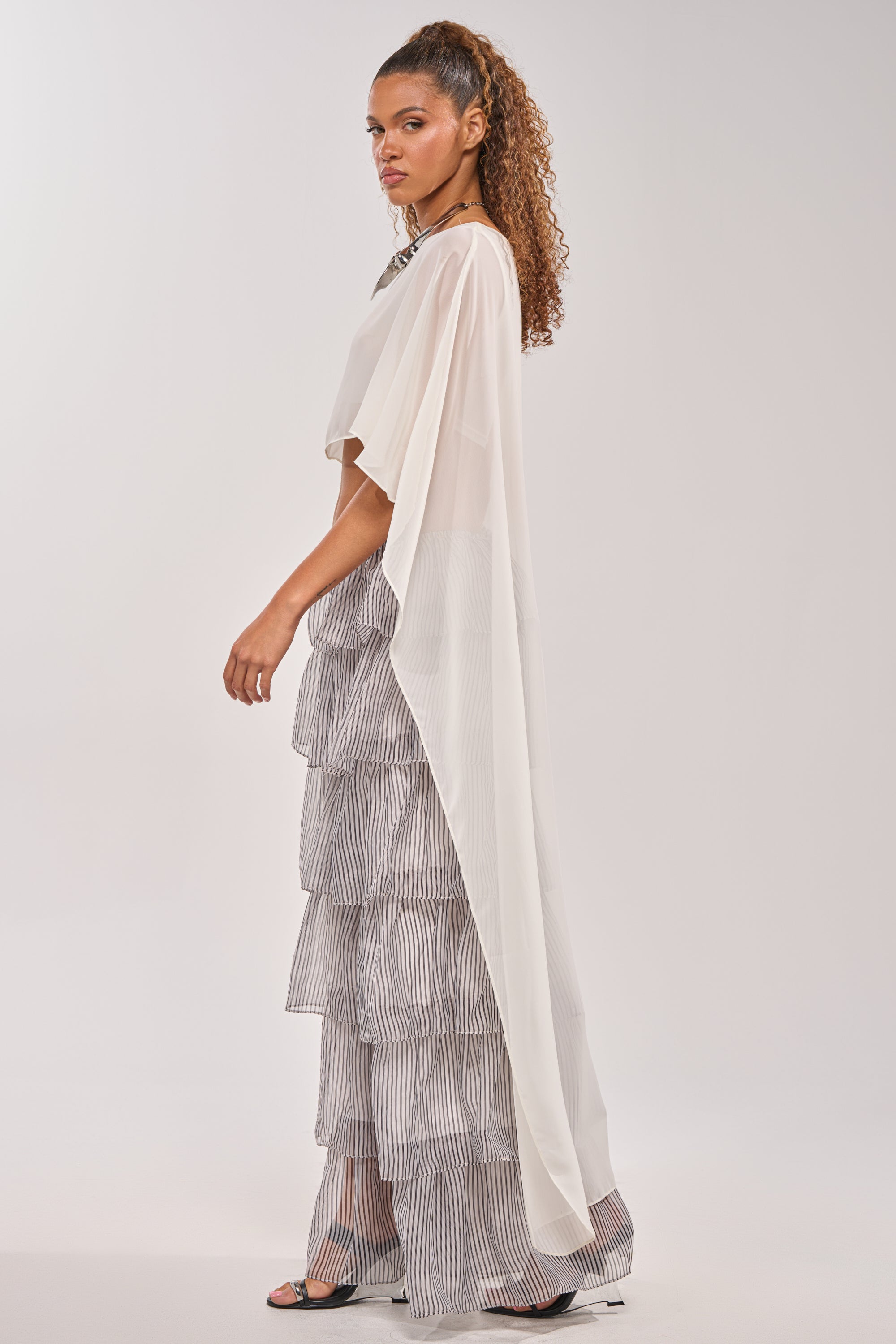 A woman stands sideways against a plain background, wearing the IT GIRL BLOUSE IN IVORY over a tiered, striped ruffle dress with a high-low hem and open-toe heels; her curly hair is styled in a high ponytail. 3