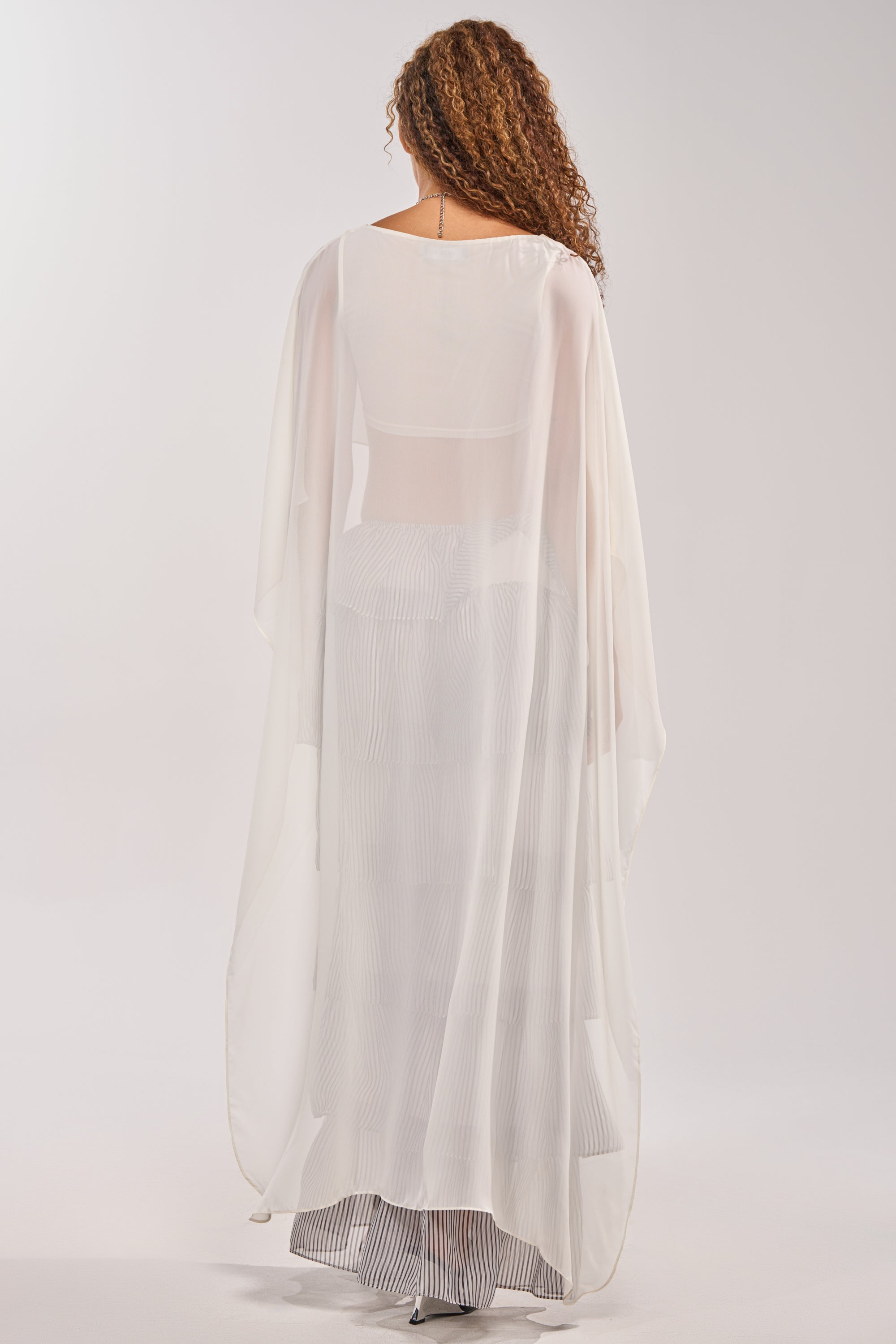 A woman with curly hair stands facing away, wearing the IT GIRL BLOUSE IN IVORY—a long, high-low chiffon cape top that brings elegance to her striped festival look against a plain, light background. 4
