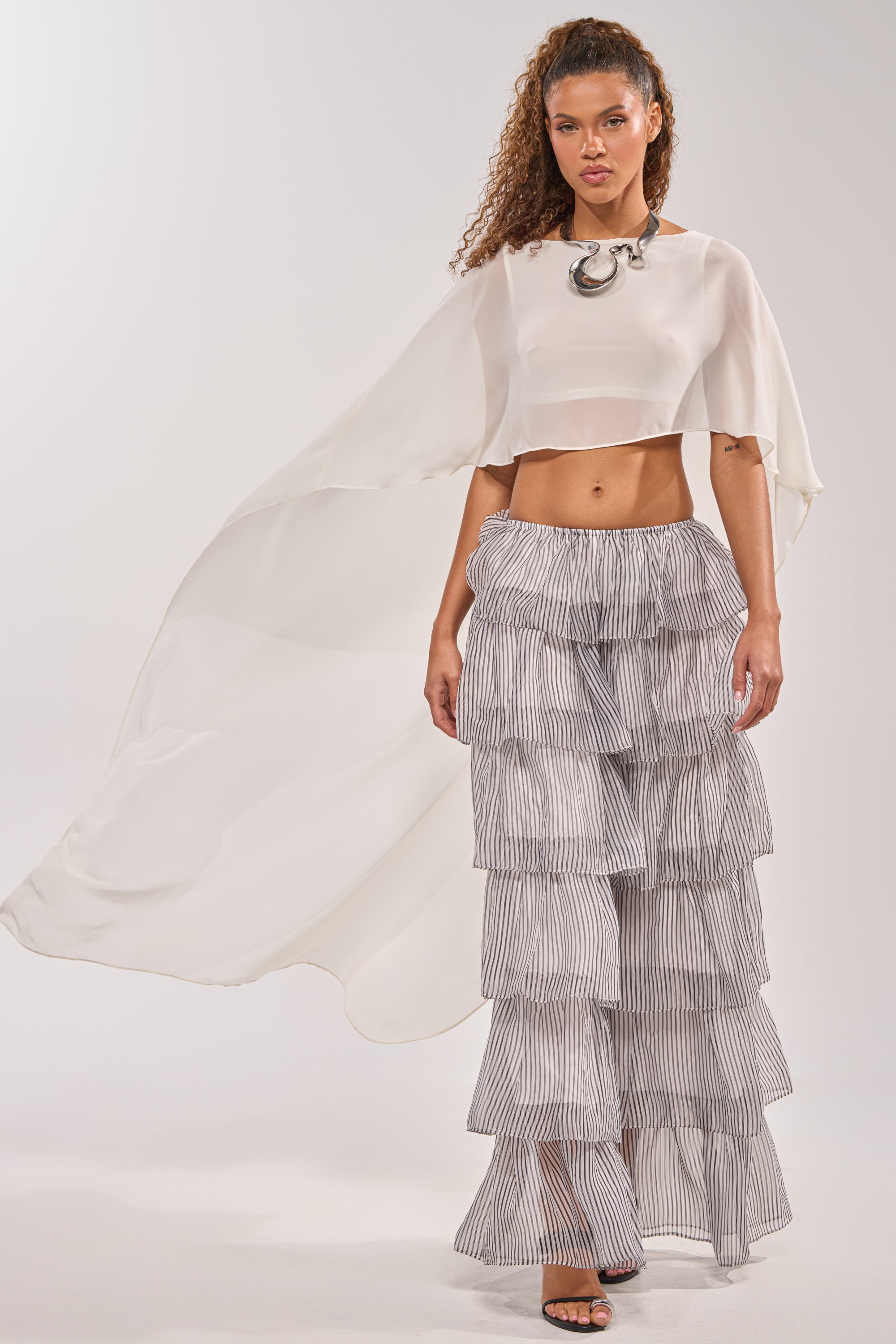 A woman exudes confidence in the IT GIRL BLOUSE IN IVORY, a bold necklace, high-waisted tiered ruffled striped pants, and black sandals—creating a stunning festival look against a plain background. 5