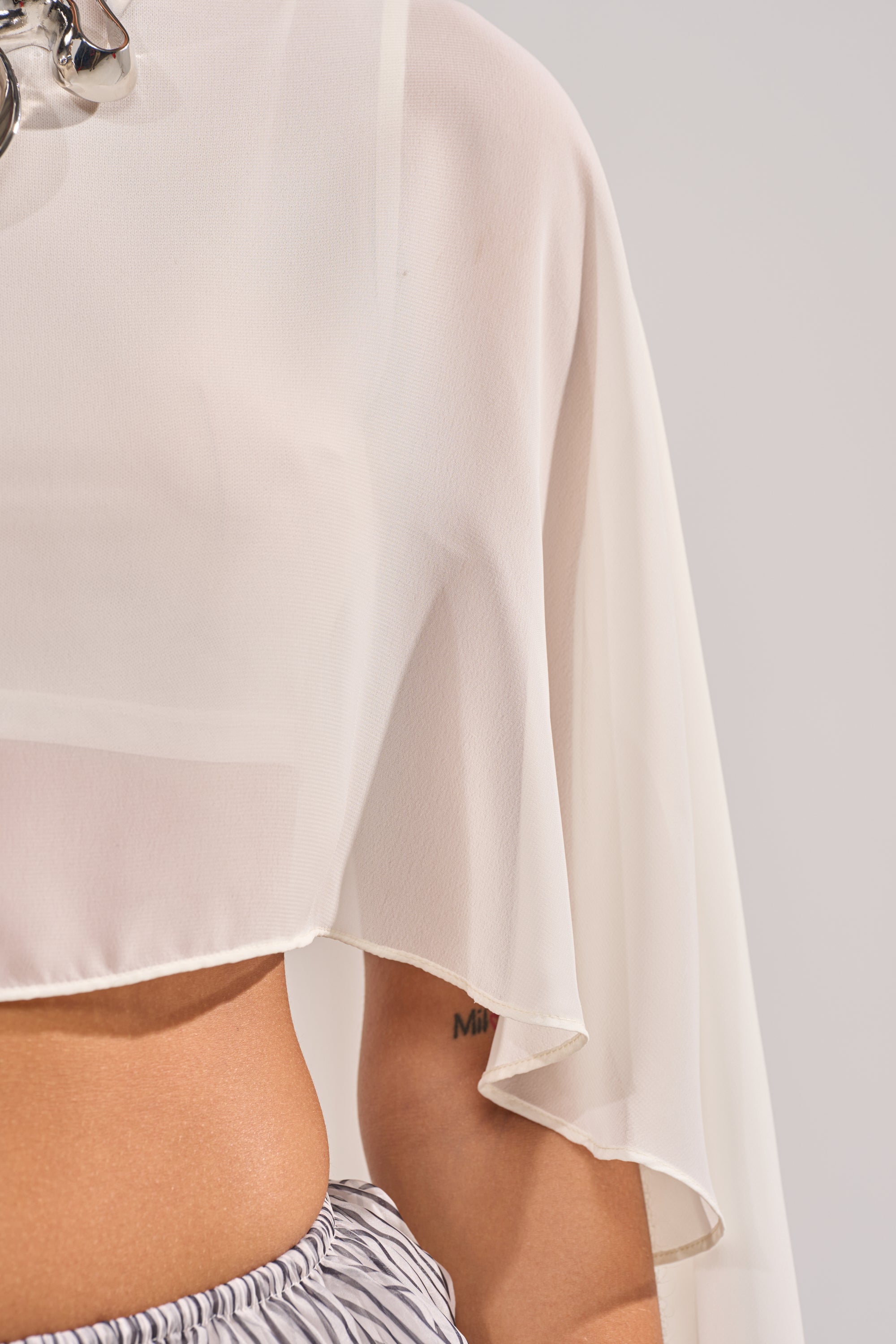 A close-up shows a person in the IT GIRL BLOUSE IN IVORY, featuring loose chiffon sleeves, paired with striped pants—an ideal festival look. Only their side and arm appear against a neutral background. 6