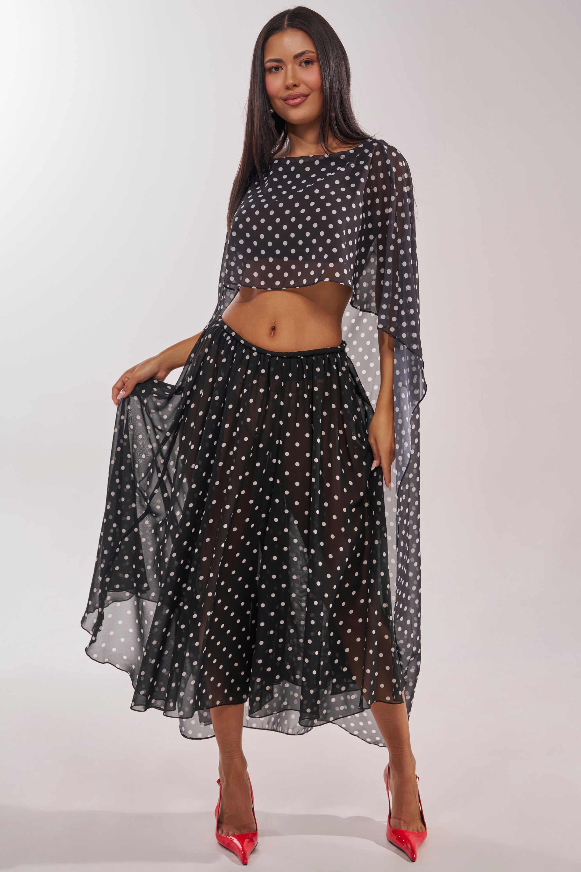 A woman showcases the IT GIRL BLOUSE, a sheer black two-piece with white polka dots and a flowing chiffon cape top, paired with a matching midi skirt and red pointed-toe heels against a simple light background. 0