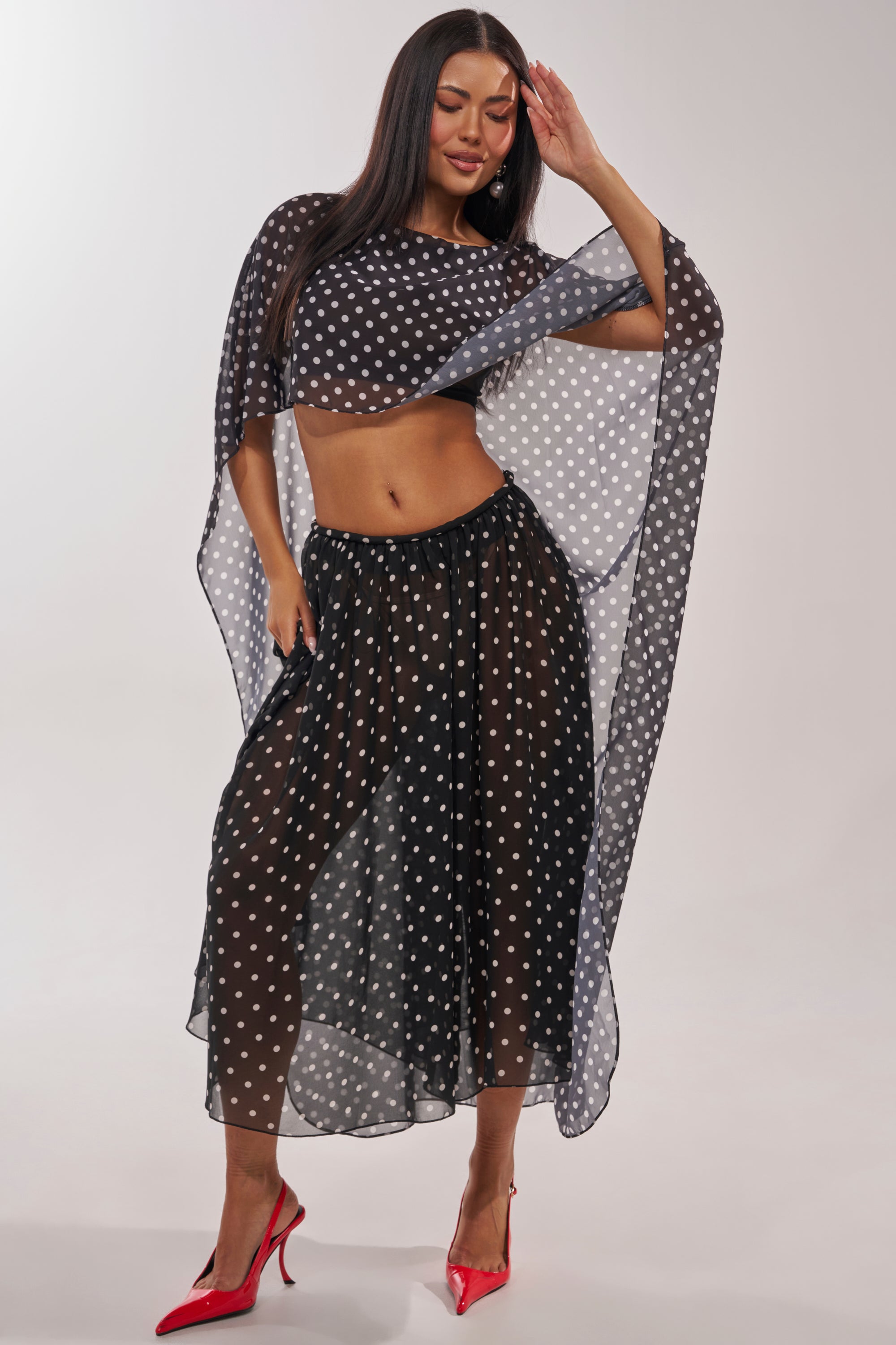 A woman exudes confidence in the IT GIRL BLOUSE, a sheer black top with white polka dots and flowy chiffon cape sleeves, paired with a matching skirt and bold red heels for a striking layered look. 1