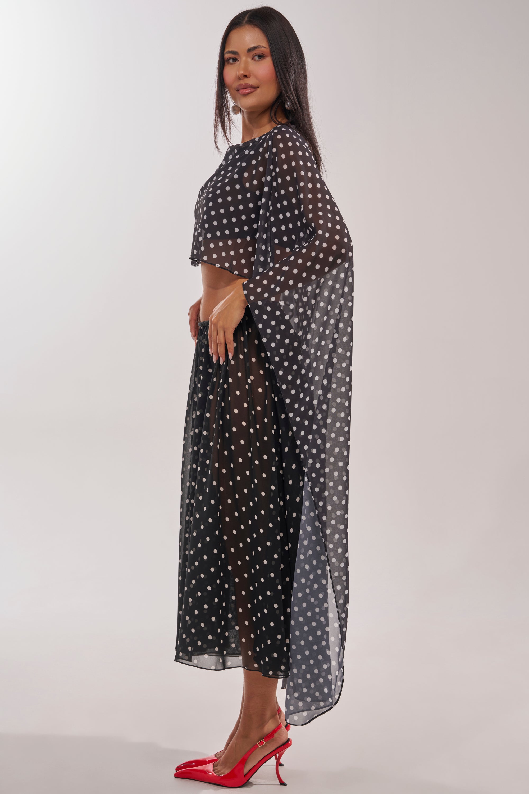 A woman with long dark hair and a neutral expression models the IT GIRL BLOUSE, a black sheer polka-dot cape top with wide sleeves, paired with cropped pants and red high heels, standing sideways against a plain background. 4