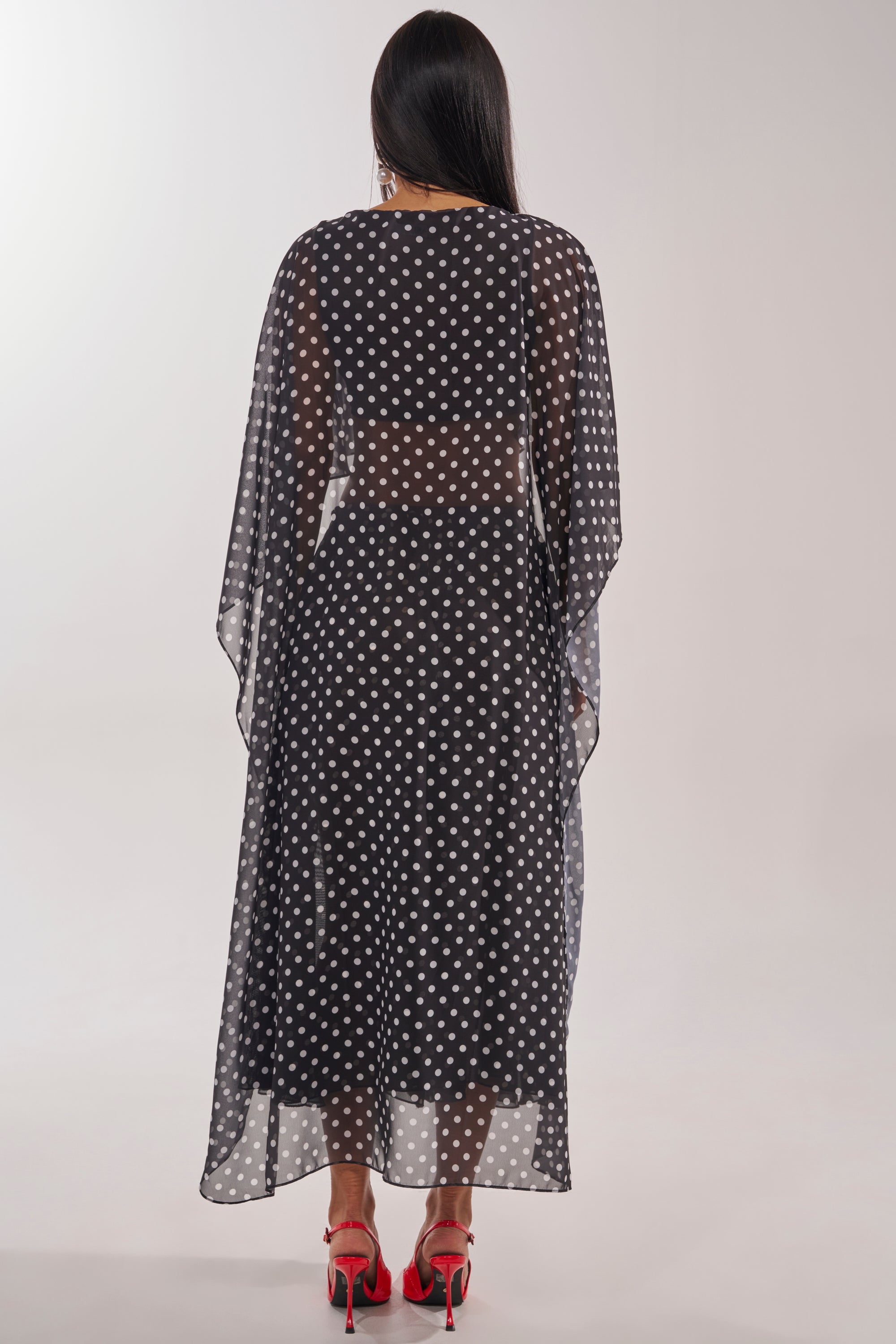 A woman faces away, wearing the IT GIRL BLOUSE—a long, sheer polka-dot chiffon cape top with a high-low silhouette—paired with red heels. Her straight dark hair flows down her back against a plain light background. 5
