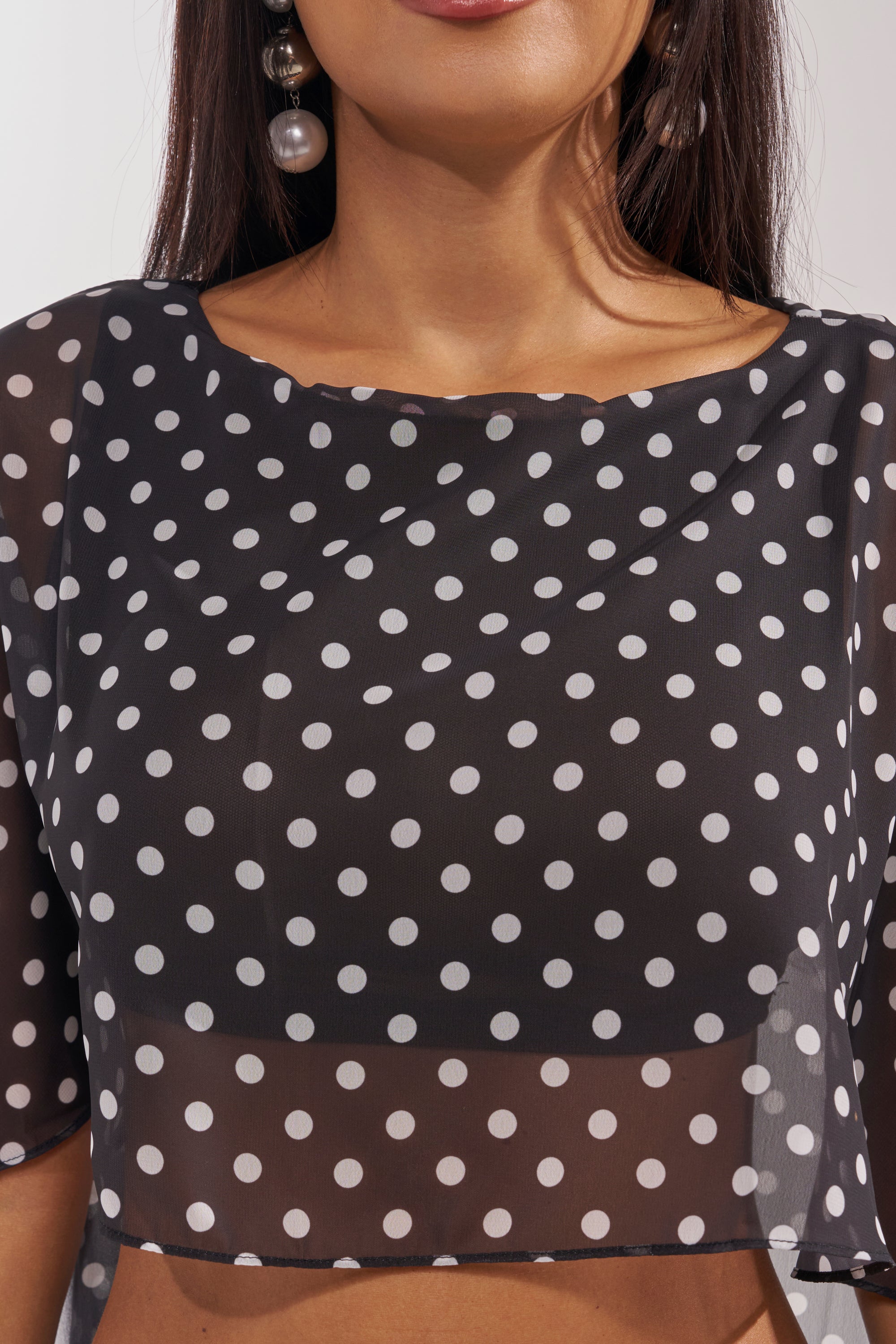 A woman wears the IT GIRL BLOUSE, a sheer black crop top with white polka dots, paired with large dangling pearl earrings. Only her lower face, neck, and upper torso are visible. 6