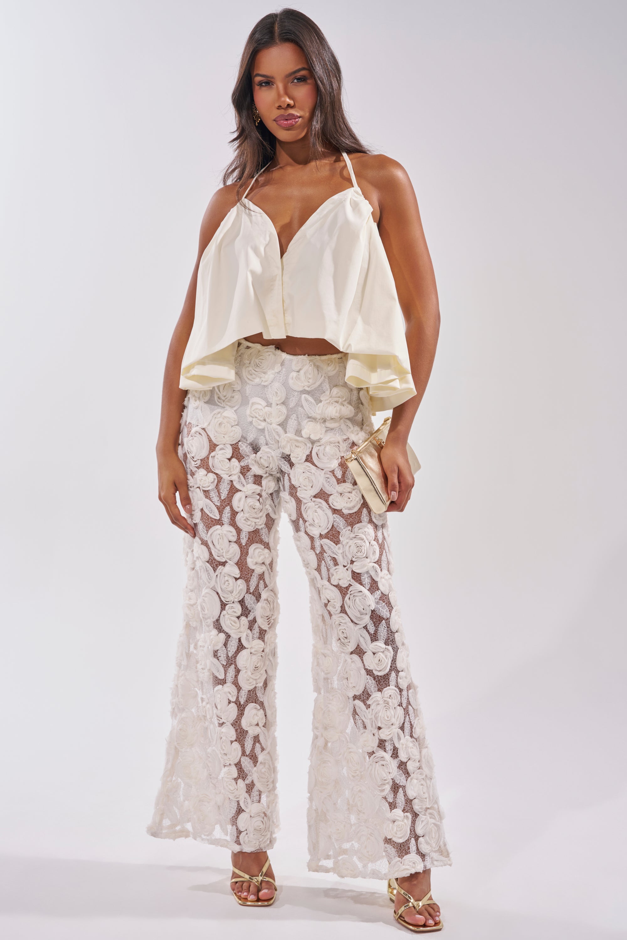 A woman wears the DINA HALTER NECK BLOUSE in ivory poplin, styled with sheer flared pants featuring white floral embroidery. She accessorizes with a beige clutch and gold heels, perfect for chic summer looks against a plain backdrop. 4