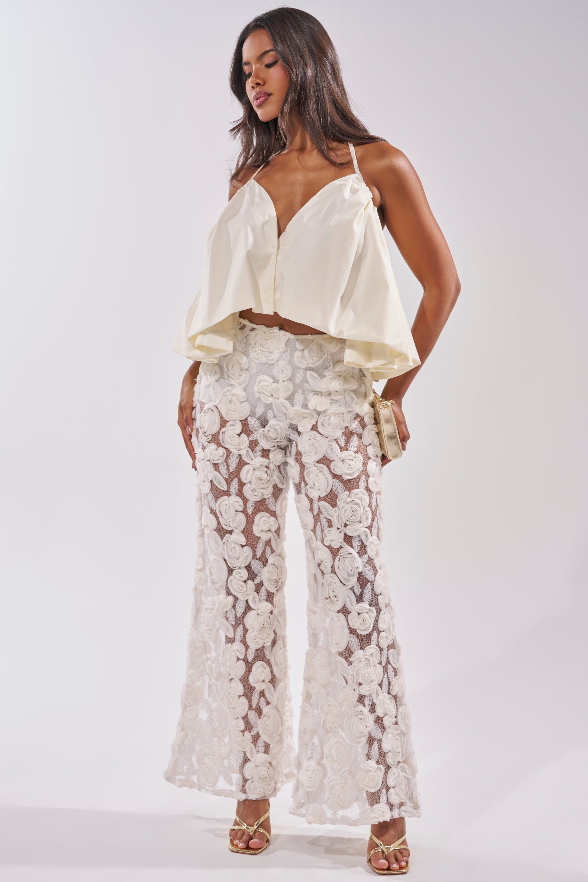 Woman in the DINA HALTER NECK BLOUSE, paired with sheer white floral lace pants, gold clutch, and strappy heels—ideal summer style against a light backdrop. 5